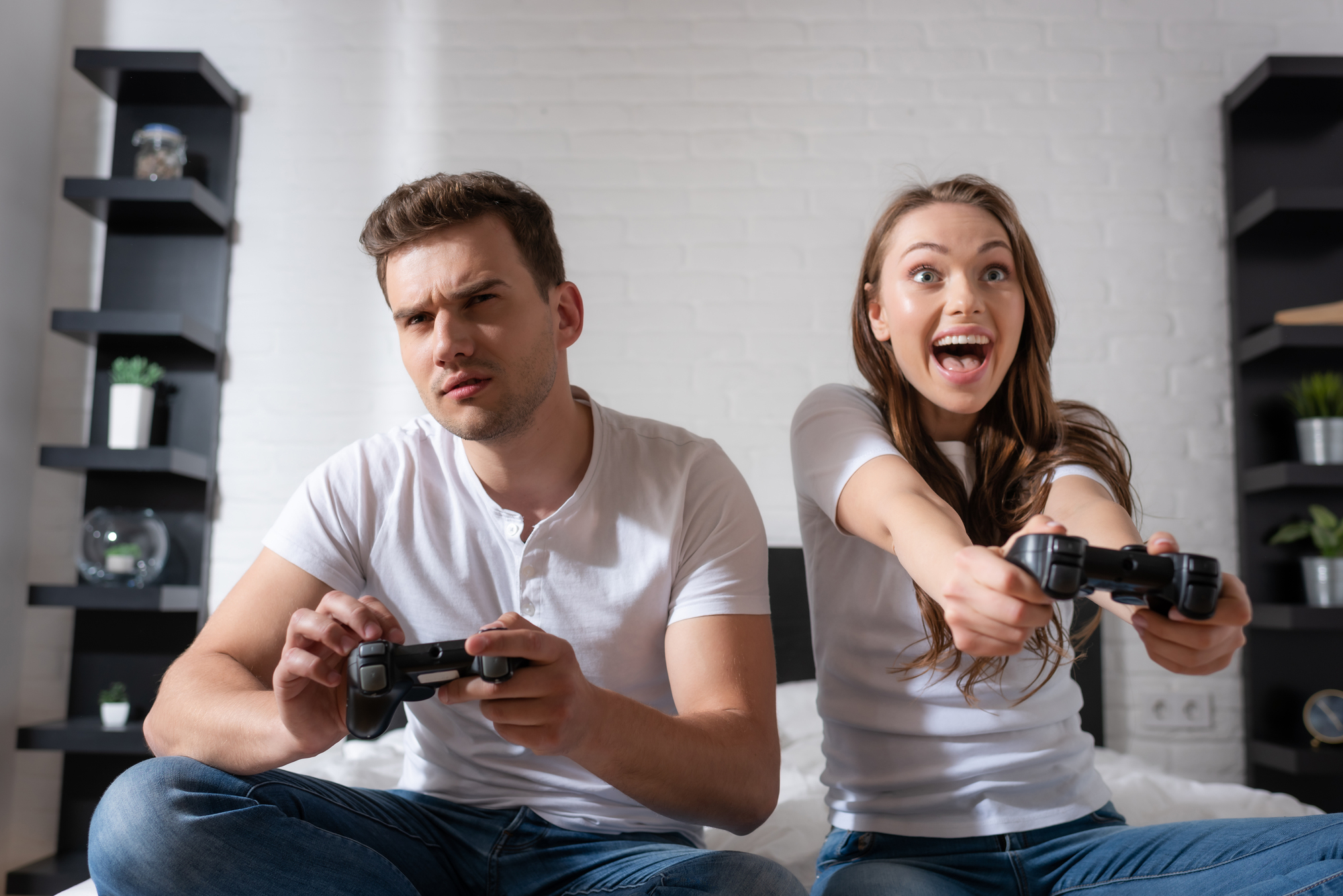 A man and a woman sit on a couch playing video games. The man appears focused while the woman looks excited. Both hold game controllers. They are in a living room with shelves and small plants in the background.