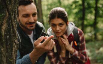 A man and woman observe a small insect on the man's fingertip while hiking in a forest. Both wear backpacks and appear intrigued by the tiny creature, surrounded by lush greenery.