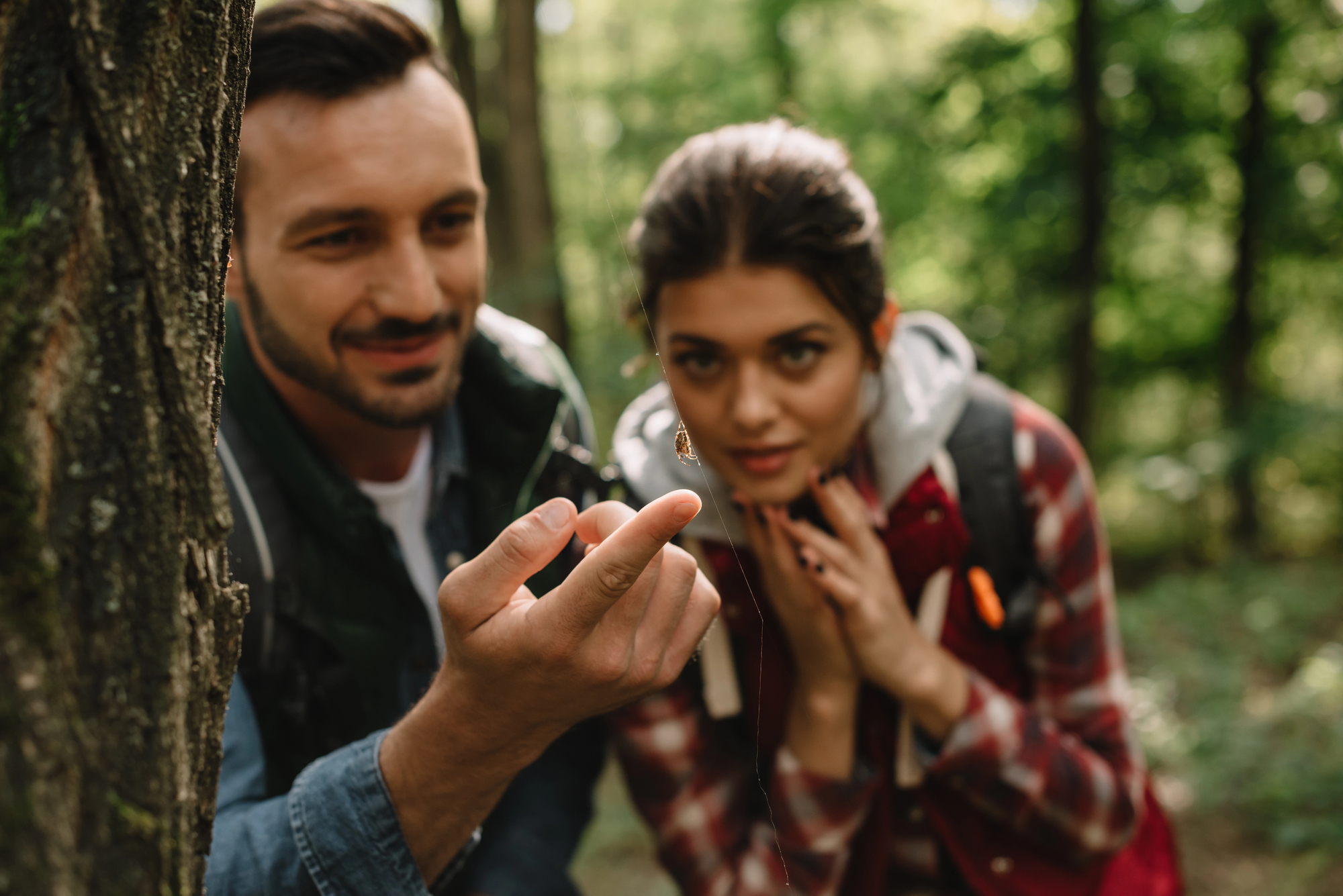 A man and woman observe a small insect on the man's fingertip while hiking in a forest. Both wear backpacks and appear intrigued by the tiny creature, surrounded by lush greenery.