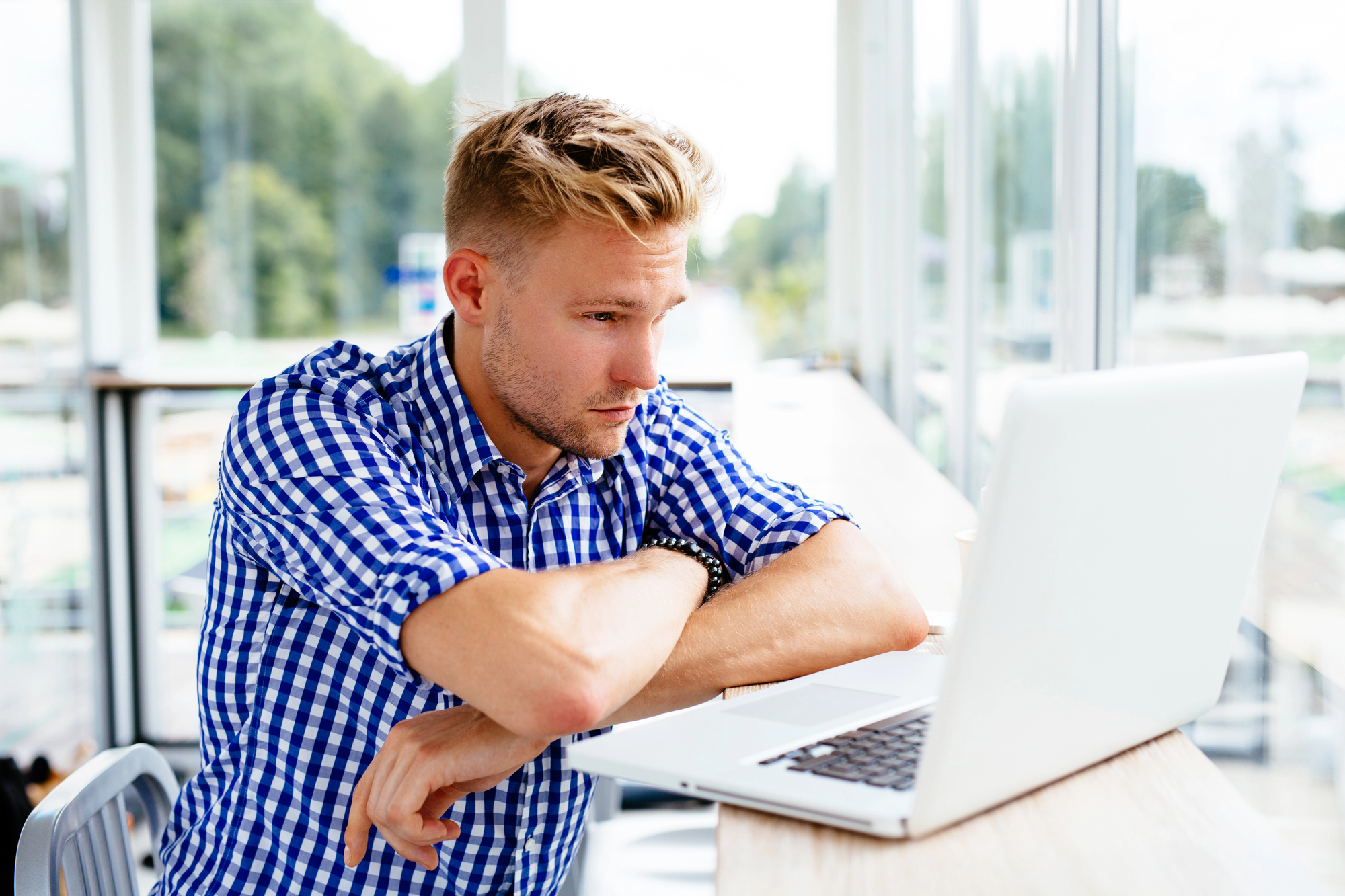 A man with short blond hair and a blue checkered shirt sits at a wooden desk, focused on a laptop in front of him. Large windows in the background reveal a blurred outdoor scene.