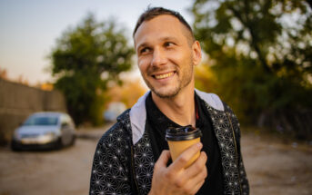 A man with short hair is smiling while holding a takeaway coffee cup outdoors. He is wearing a black jacket with a geometric pattern on the sleeves. The background shows a car parked on a dirt road and trees with green and yellow leaves.