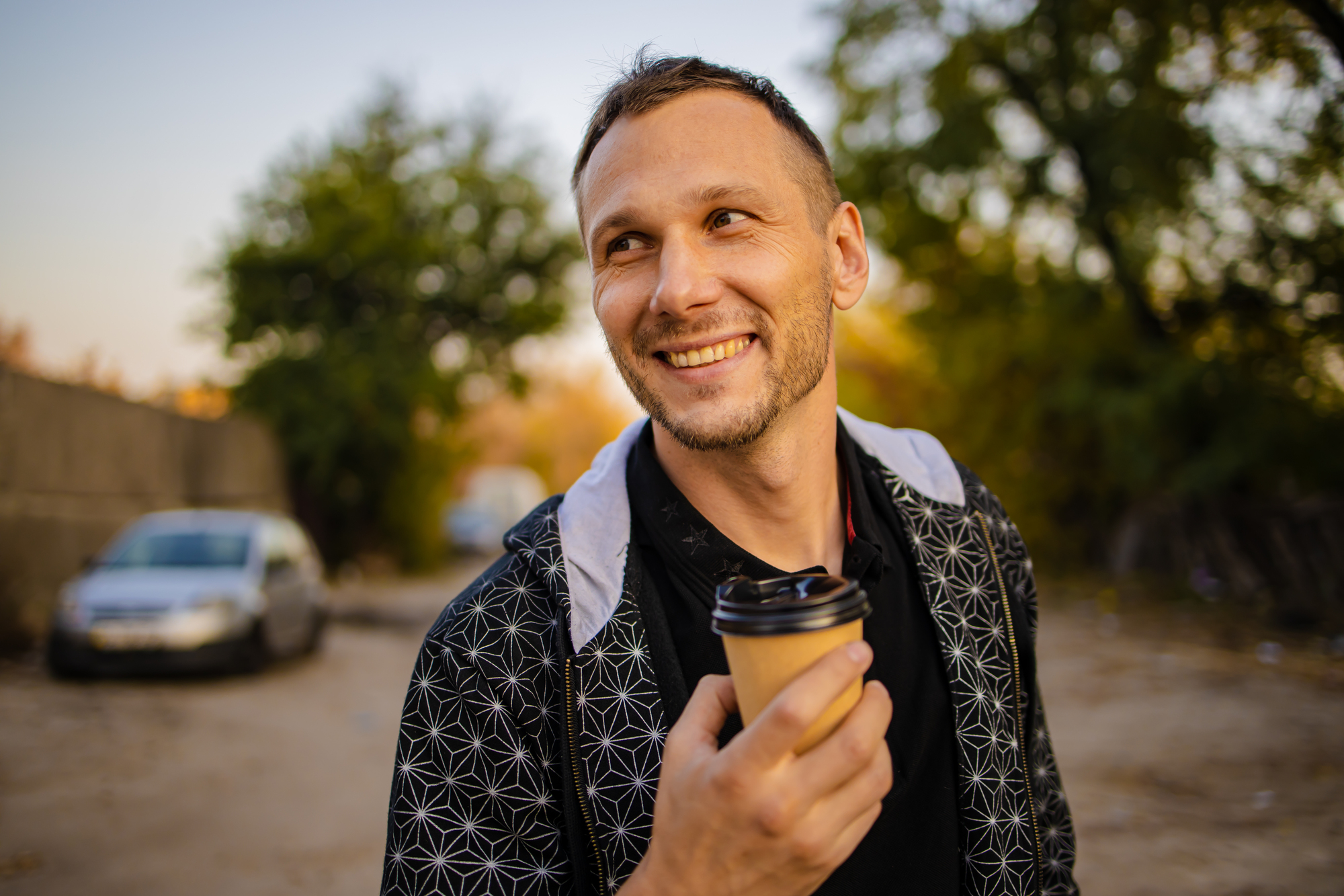 A man with short hair is smiling while holding a takeaway coffee cup outdoors. He is wearing a black jacket with a geometric pattern on the sleeves. The background shows a car parked on a dirt road and trees with green and yellow leaves.