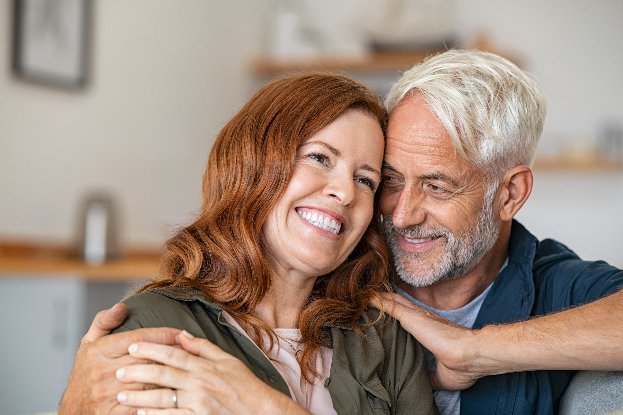 A smiling couple is sitting closely together. The older man with white hair is embracing the woman with long red hair, who is leaning into him. They both look happy and content, sitting in a cozy indoor setting.