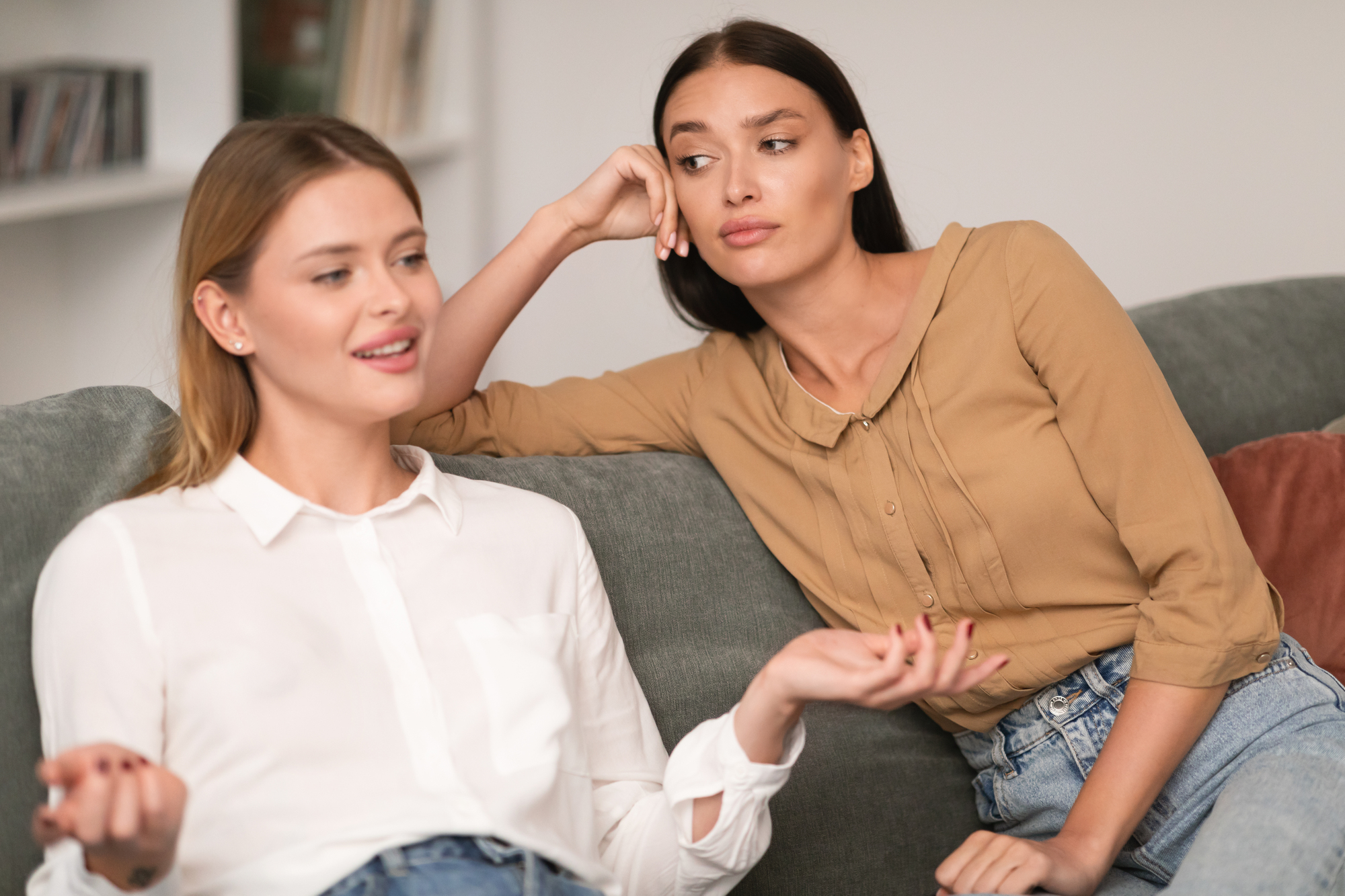 Two women sitting on a couch in a living room. The woman on the left, wearing a white shirt, is speaking while gesturing with her hands. The woman on the right, in a brown blouse, leans her head on her hand, listening intently.