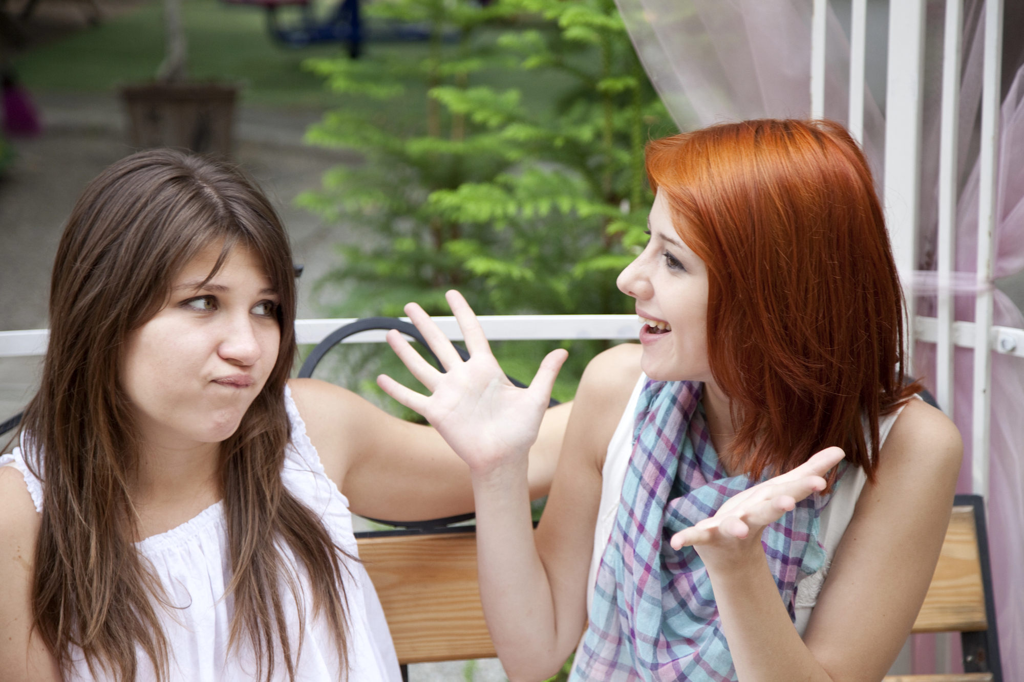 Two women sitting outside on a bench. The woman on the right with red hair is smiling and gesturing with her hands, while the woman on the left with brown hair appears unimpressed, looking away with a slight smirk.