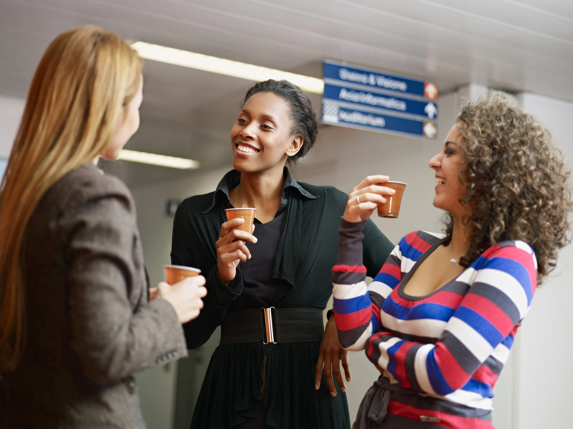 Three people enjoying a casual conversation while holding coffee cups in a bright indoor setting. They are smiling and standing near a hallway with signs above them.