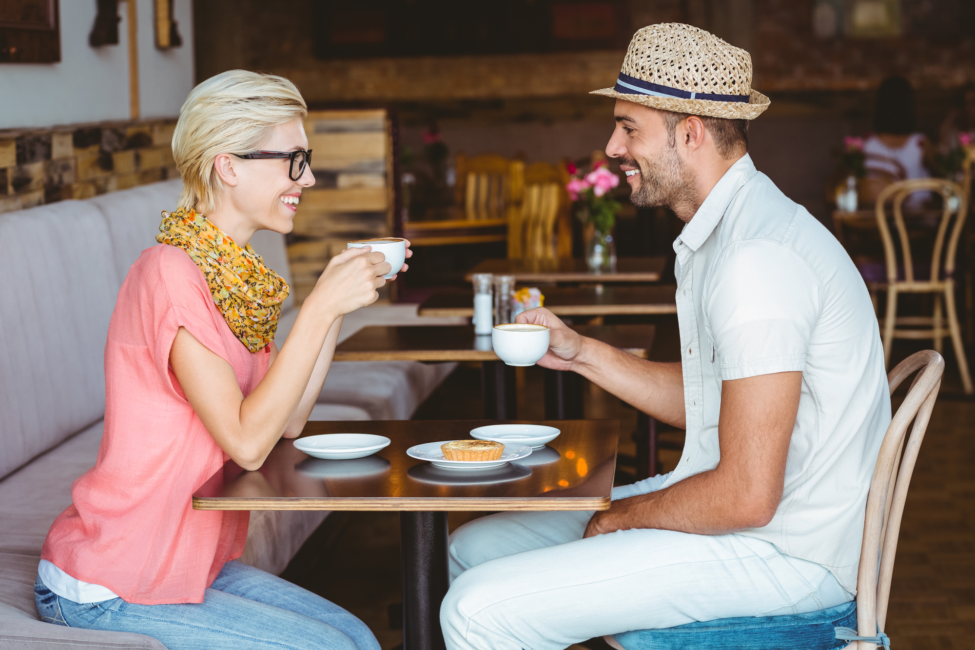 A man and woman smile at each other while sitting at a café table, each holding a cup. The woman wears glasses and a scarf, and the man wears a hat. There's a pastry on the table in a cozy, warmly lit setting.