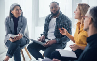 A diverse group of four people sit together in a circle, engaged in discussion. One person holds a notebook, another a laptop. The setting is a well-lit room with soft natural light coming through large windows.