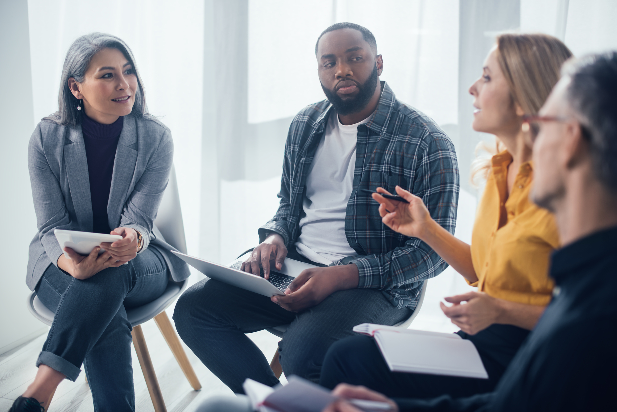 A diverse group of four people sit together in a circle, engaged in discussion. One person holds a notebook, another a laptop. The setting is a well-lit room with soft natural light coming through large windows.