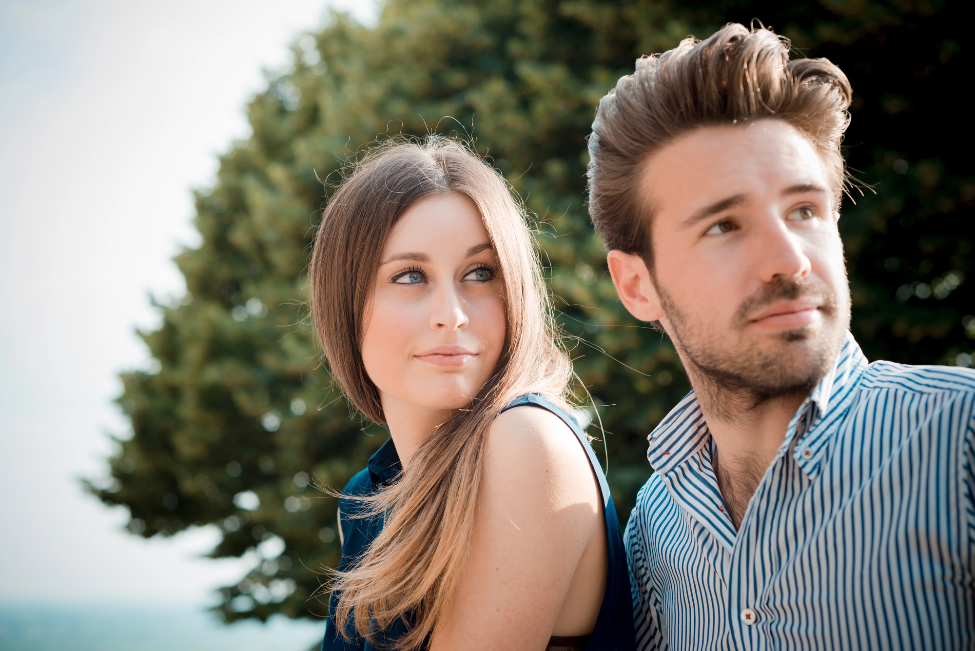 A woman and man sitting outdoors, both gazing into the distance. The woman has long hair, wearing a sleeveless top, while the man wears a striped shirt. A tree is visible in the background.