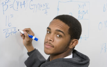 A young man stands in front of a whiteboard filled with mathematical equations, holding a blue marker. He wears a gray hoodie and looks over his shoulder towards the camera. The whiteboard shows various algebraic expressions and graphs.