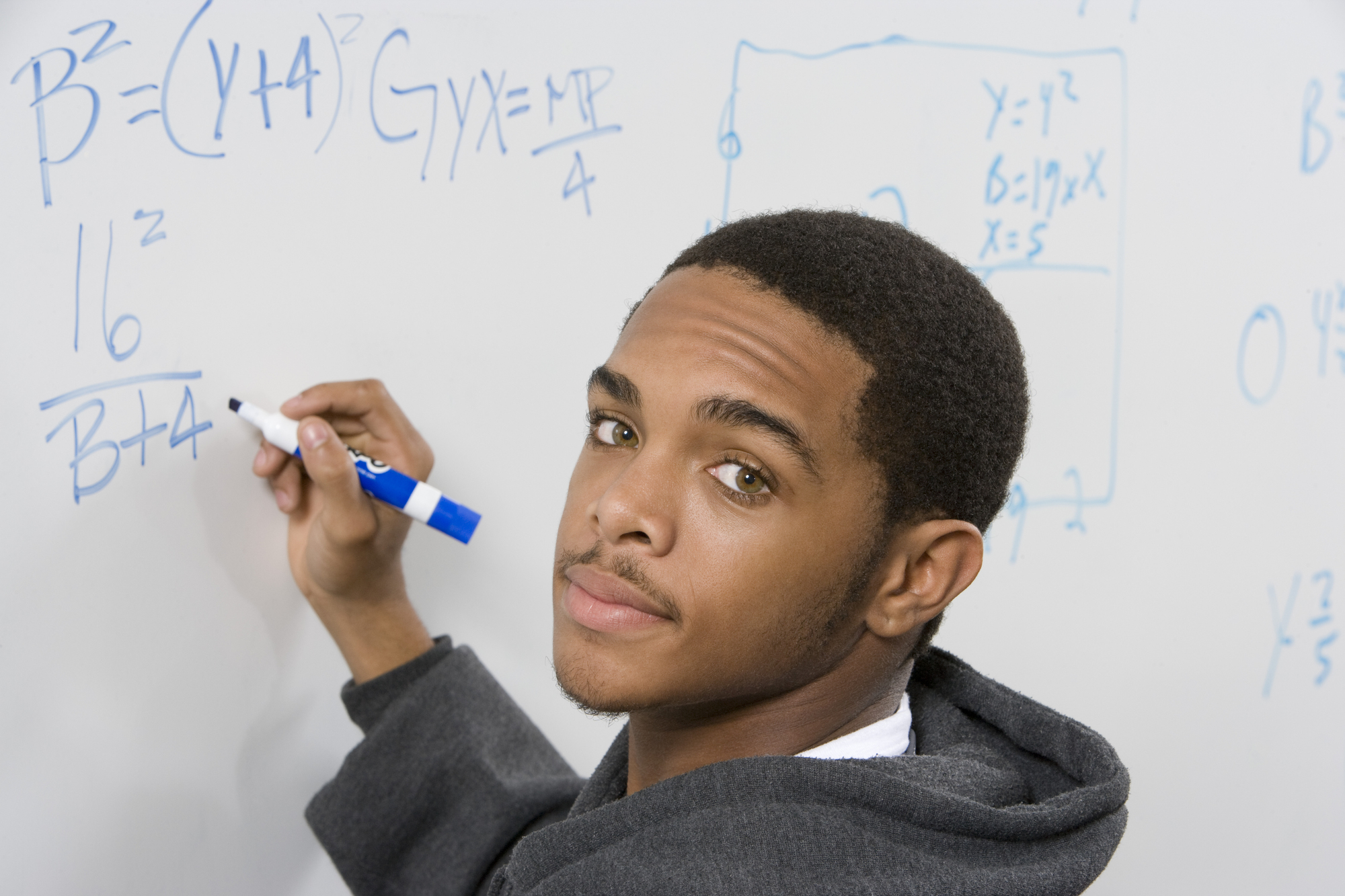 A young man stands in front of a whiteboard filled with mathematical equations, holding a blue marker. He wears a gray hoodie and looks over his shoulder towards the camera. The whiteboard shows various algebraic expressions and graphs.