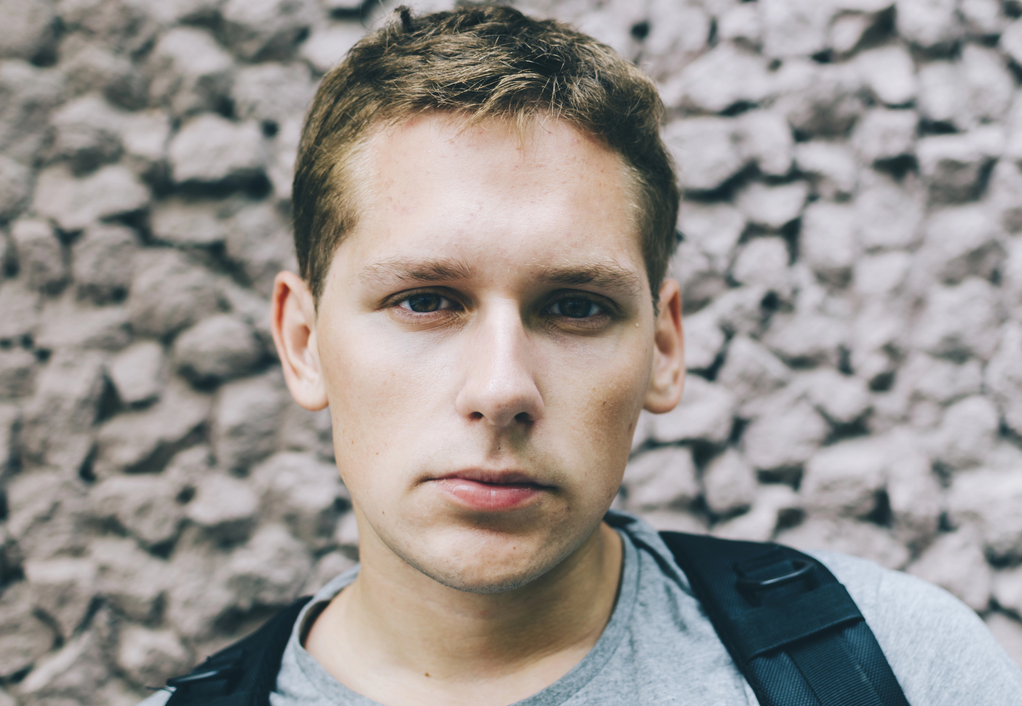 A young person with short brown hair and a serious expression stands in front of a textured stone wall, wearing a gray shirt and a black backpack.