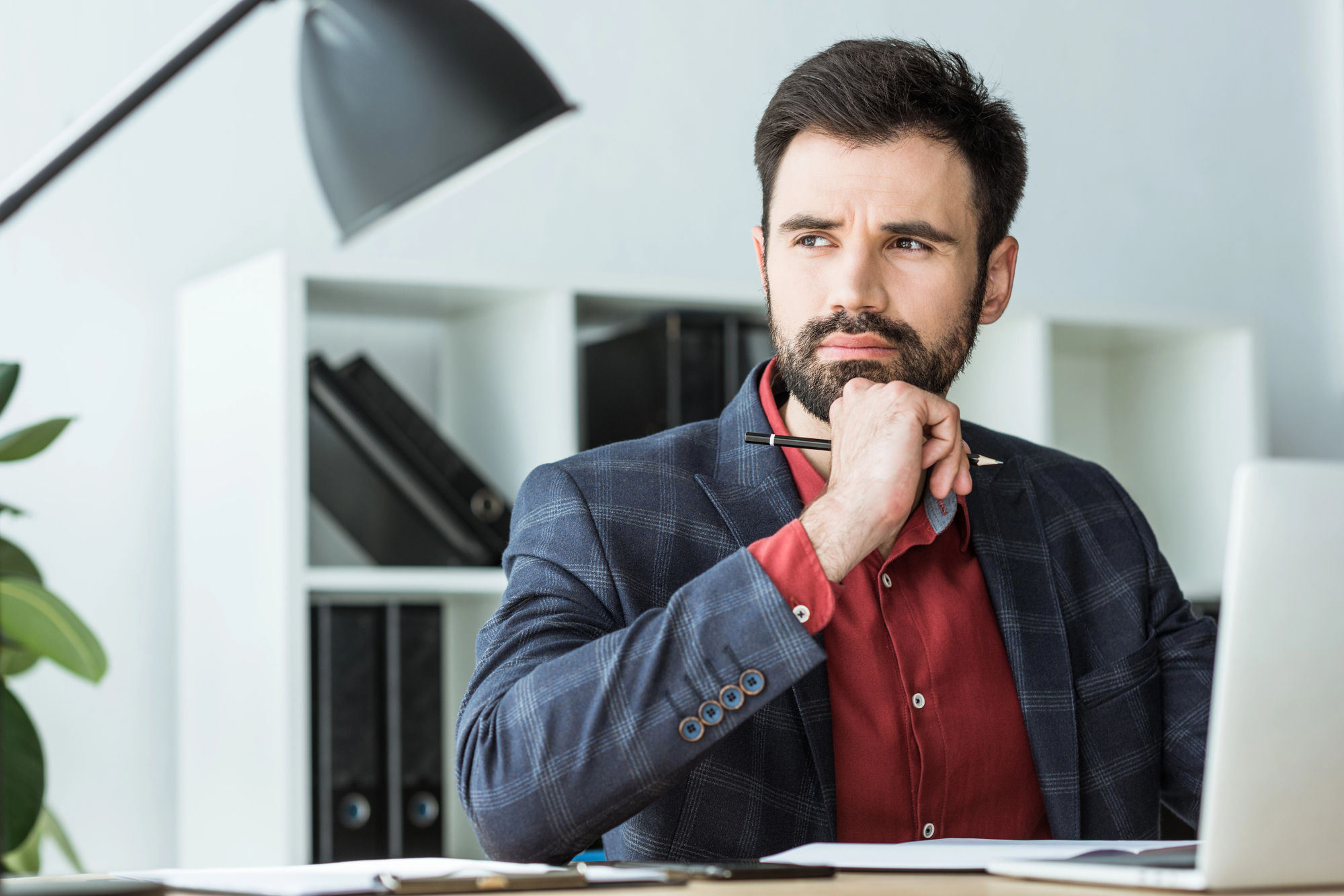 A man with a beard sits at a desk, holding a pen to his chin in a thoughtful pose. He wears a red shirt and a plaid blazer. Behind him are shelves with books and a lamp. A laptop is open in front of him.