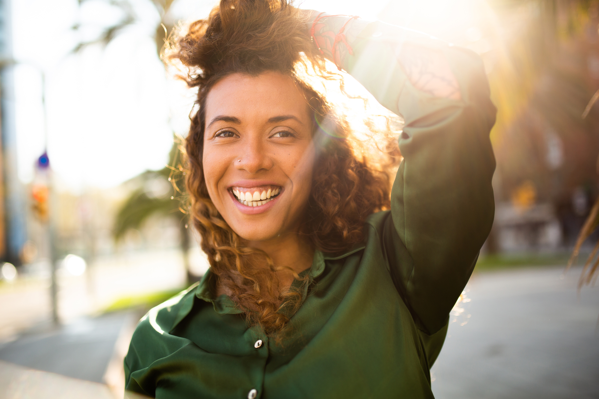 A woman with curly hair smiles brightly while standing outdoors in sunlight. She is wearing a green shirt, and her hand is raised to her head. The background is blurred with hints of trees and buildings.