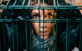 A woman with curly hair looks through the bars of a metal fence. She wears black gloves, a scarf, and has an intense expression. The focus is on her face and eyes, conveying a sense of entrapment or curiosity.