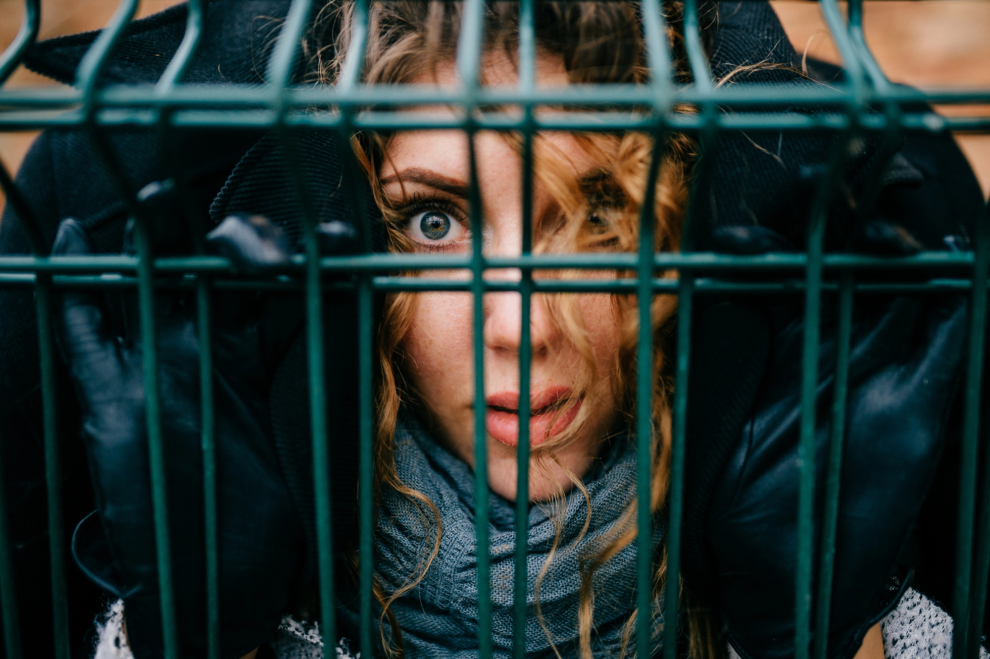 A woman with curly hair looks through the bars of a metal fence. She wears black gloves, a scarf, and has an intense expression. The focus is on her face and eyes, conveying a sense of entrapment or curiosity.