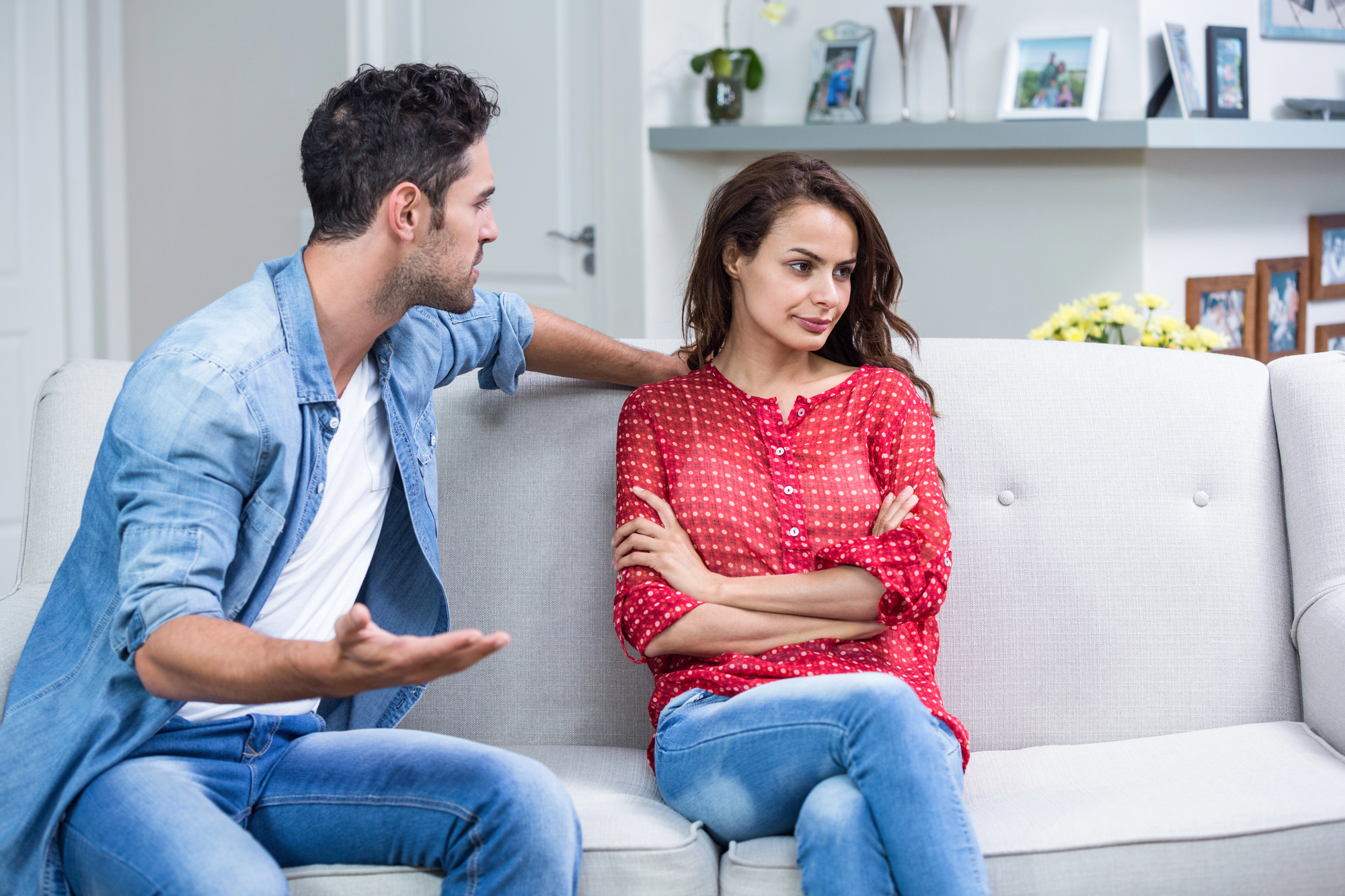 A man and woman are sitting on a couch in a living room. The man, wearing a denim shirt, appears to be explaining something with an open hand gesture. The woman, in a red polka-dot blouse, is looking away with her arms crossed.