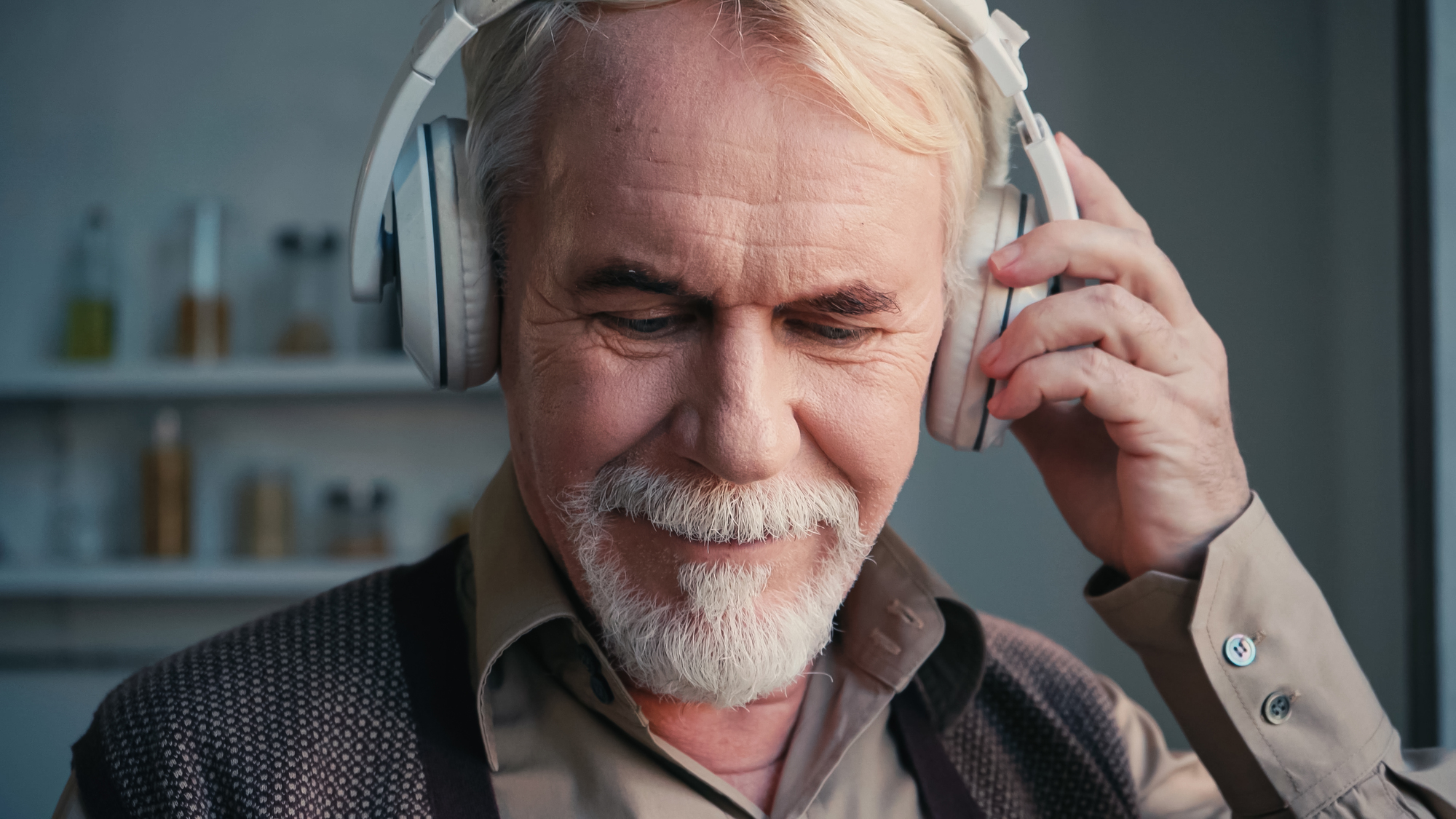 An older man with a beard and white hair is smiling while wearing headphones. He holds one hand up to his ear, appearing to enjoy listening to music. He is dressed in a collared shirt and sweater, with a blurred background.