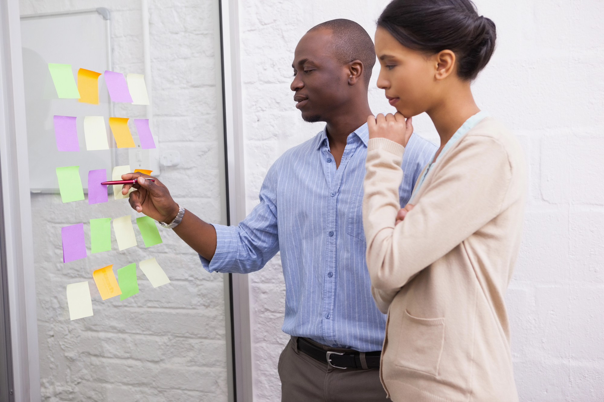 Two people are standing by a glass board with colorful sticky notes. One person is pointing at a note with a pen, while the other observes with a thoughtful expression. They appear to be collaborating in a bright office setting.