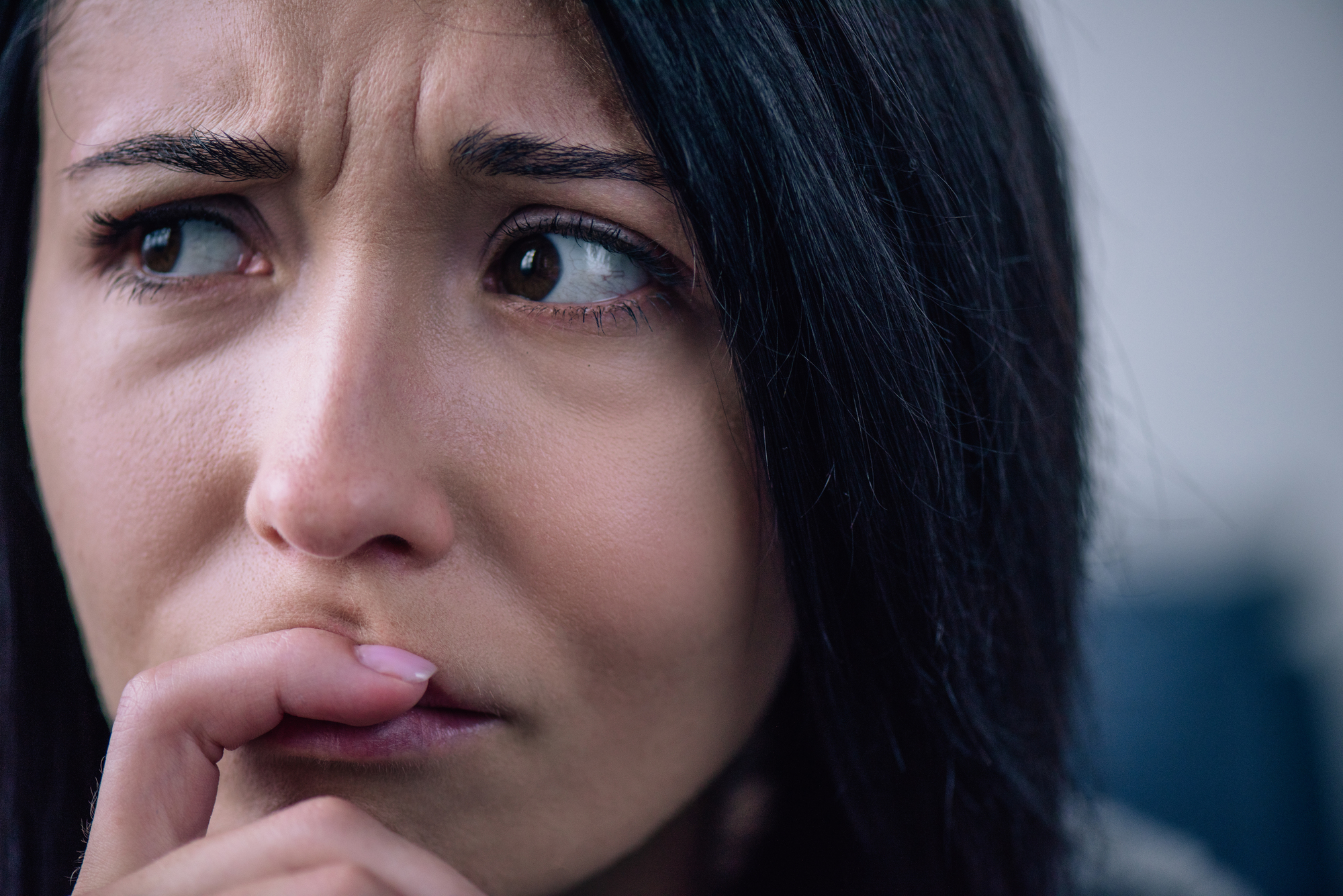 Close-up of a worried-looking woman with her hand near her mouth. Her eyes appear to be looking to the side, and her eyebrows are furrowed, suggesting concern or anxiety. She has dark hair and smooth skin, with a neutral background.