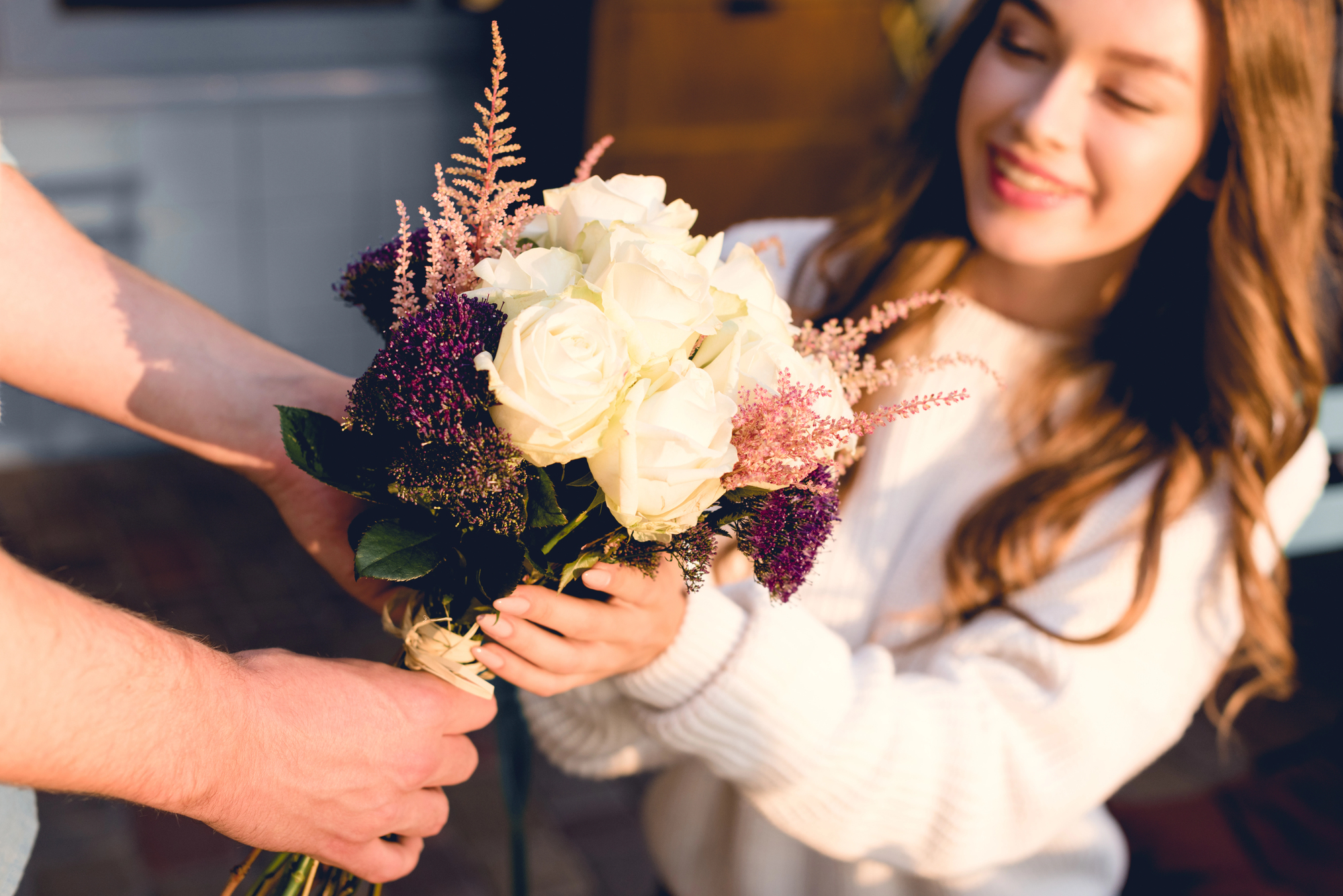 A person with long hair, wearing a white sweater, smiles while receiving a bouquet of white and purple flowers from another person. The scene is warmly lit, creating a cheerful and pleasant atmosphere.