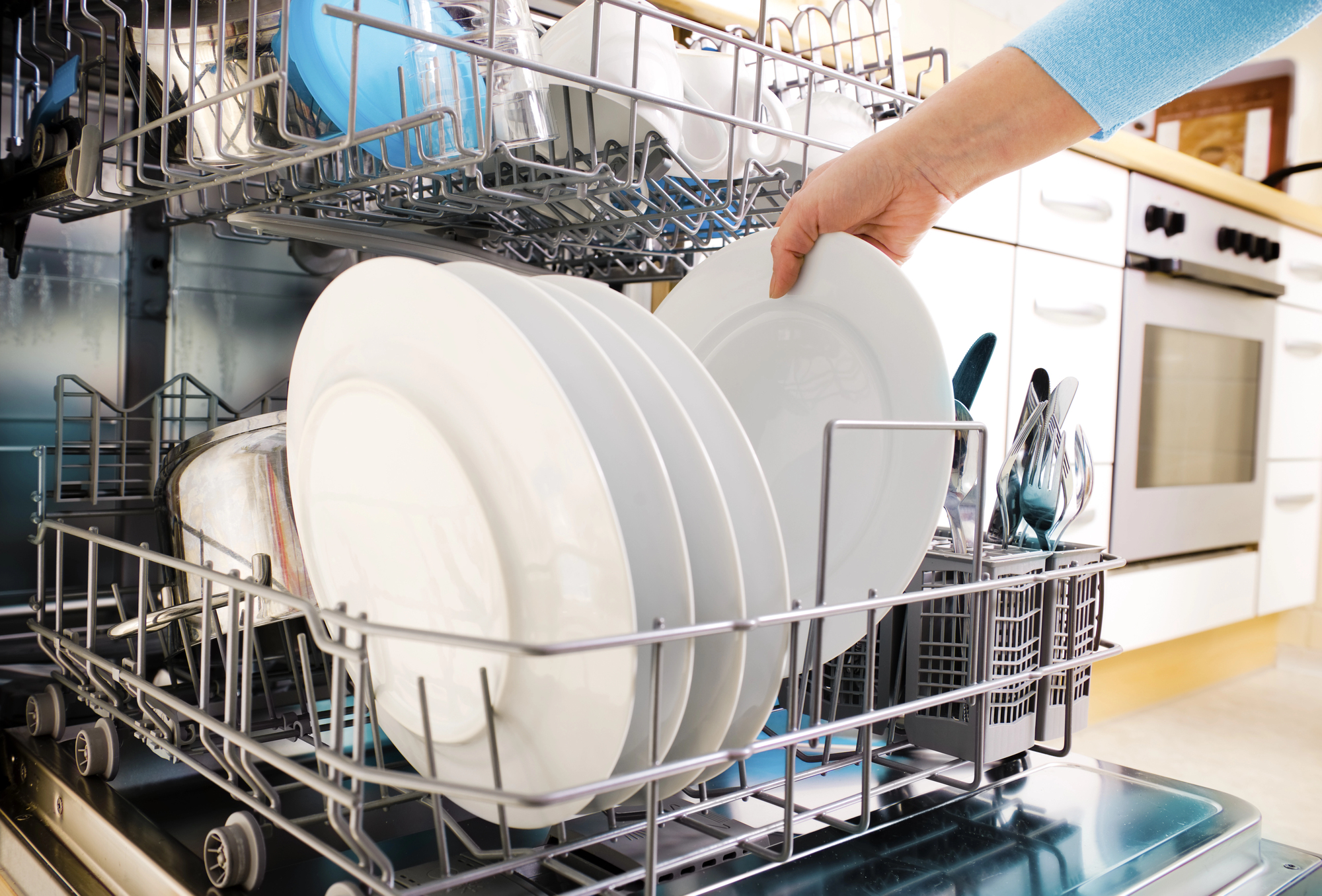 A person placing a white plate into the lower rack of an open dishwasher, which contains other clean dishes and cutlery. Kitchen cabinets and countertops are visible in the background.