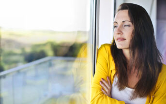 A woman in a yellow jacket stands by a window, gazing thoughtfully outside. Her arms are crossed, and her reflection is visible on the glass. The background shows a blurred view of greenery.