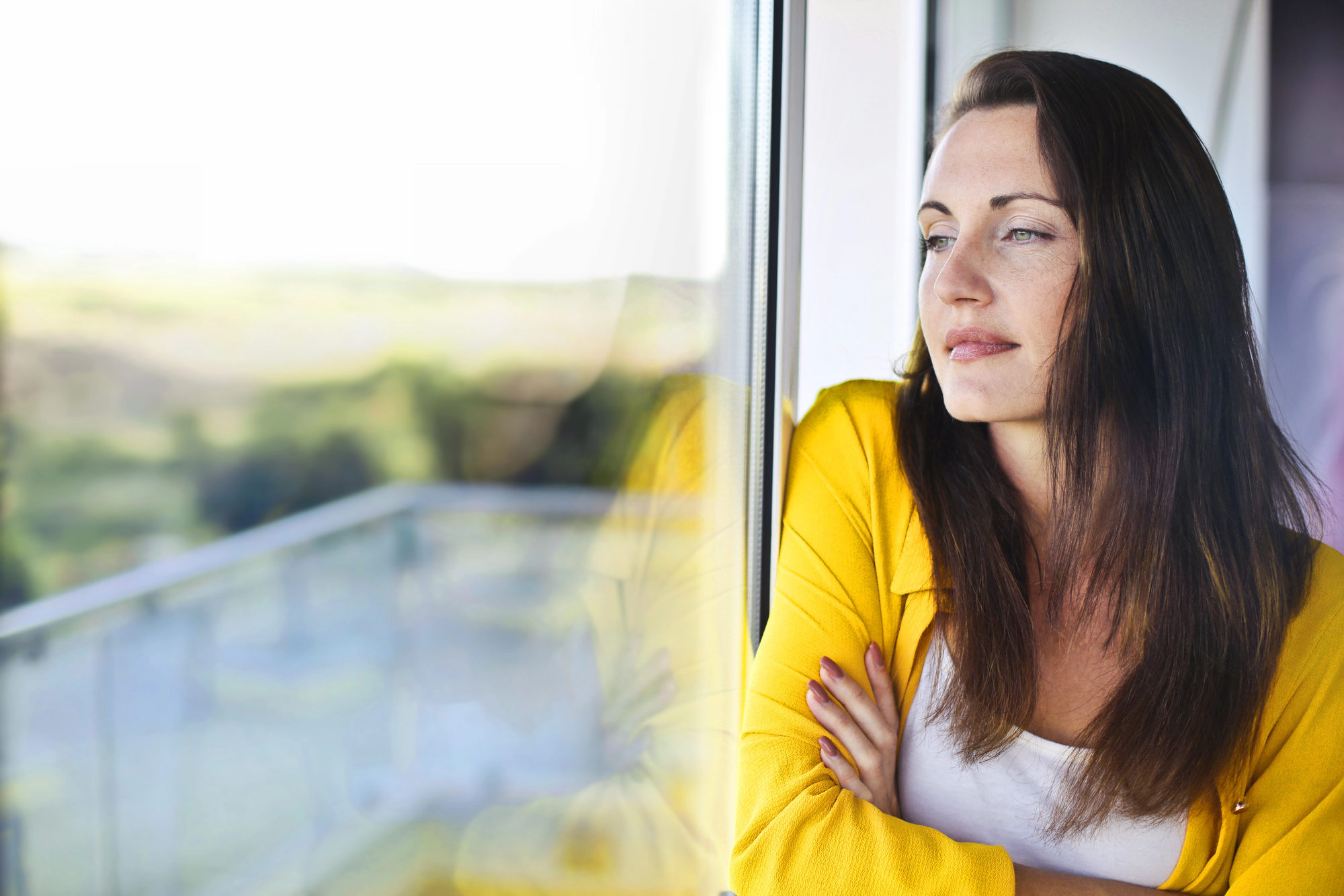 A woman in a yellow jacket stands by a window, gazing thoughtfully outside. Her arms are crossed, and her reflection is visible on the glass. The background shows a blurred view of greenery.