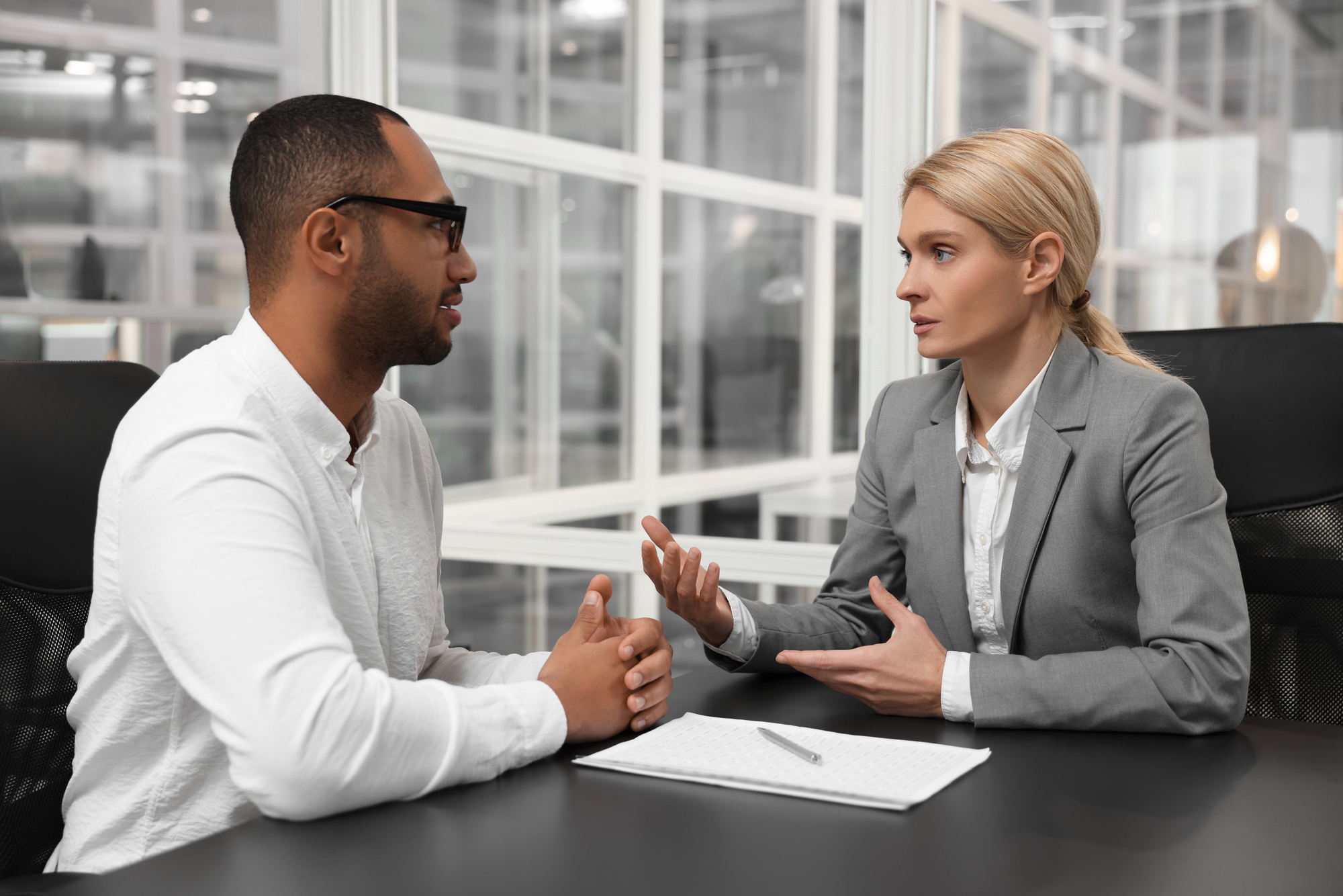 A man and a woman are having a serious discussion in an office. The man is wearing a white shirt and glasses, and the woman is in a gray suit. A notebook and pen are on the table between them.