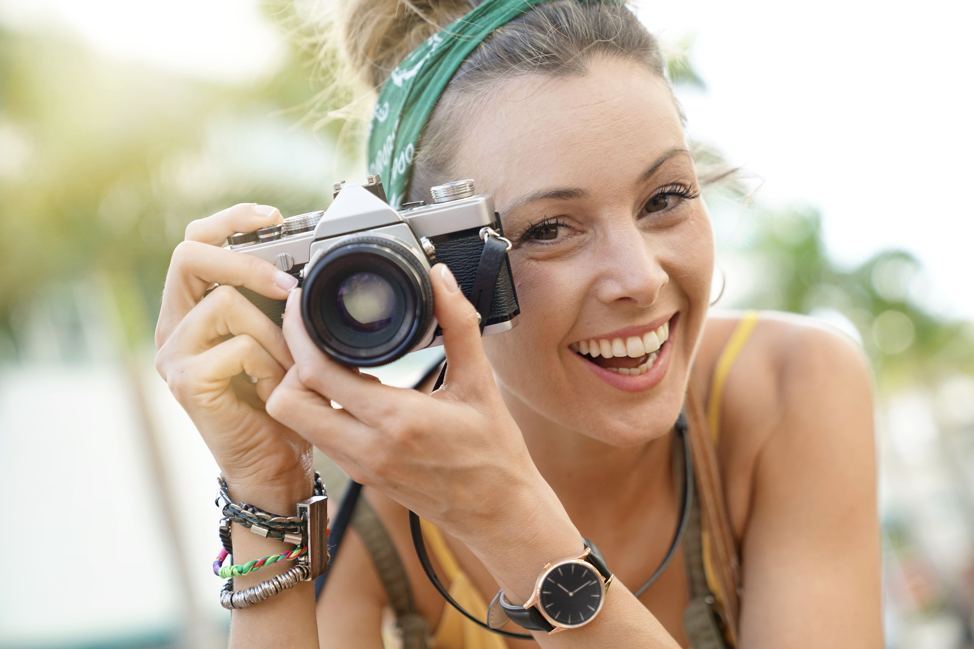 A smiling woman with a green headband and a wristwatch holds a retro camera outdoors. She wears a yellow top and bracelets, and palm trees are blurred in the background.