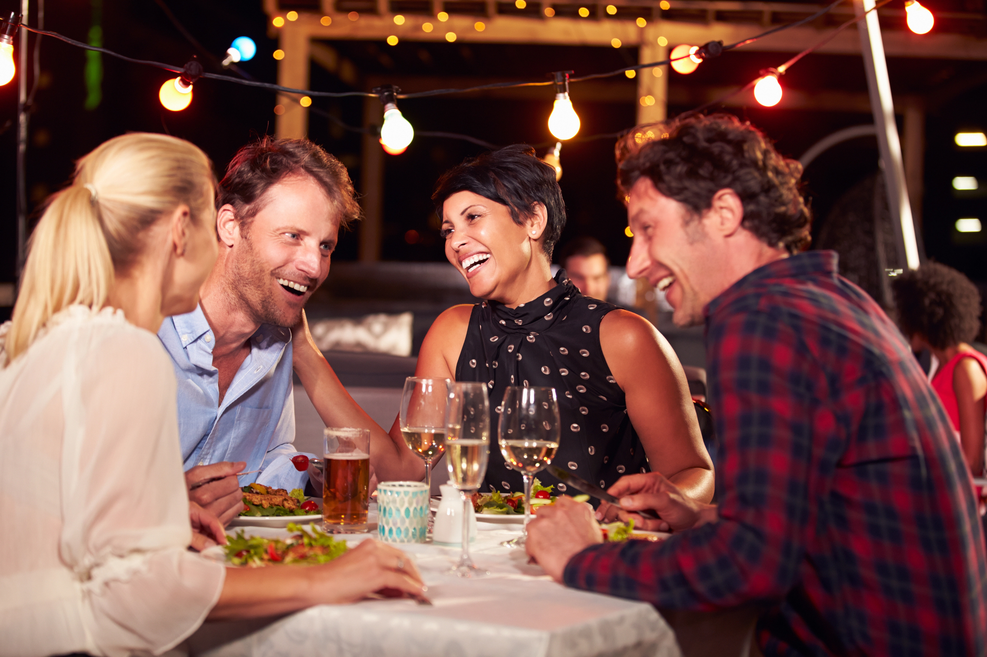 Four people are gathered around a table at an outdoor restaurant, enjoying a meal together. They are smiling and laughing, with plates of food and glasses of wine and beer in front of them. String lights create a warm atmosphere.