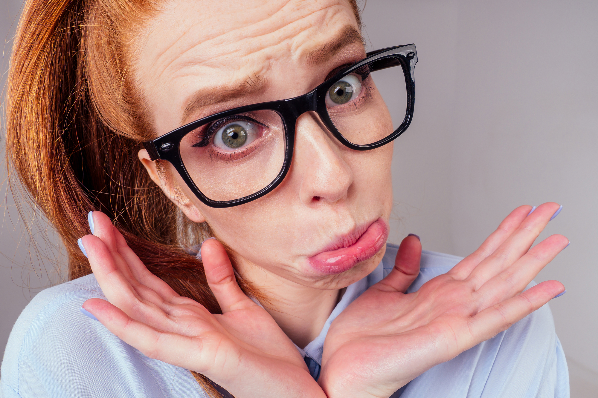 A person with long red hair and black-framed glasses is making a pouty face with wide-open eyes. They hold their hands up near their chin, palms facing outward. They are wearing a light blue shirt and are set against a plain background.