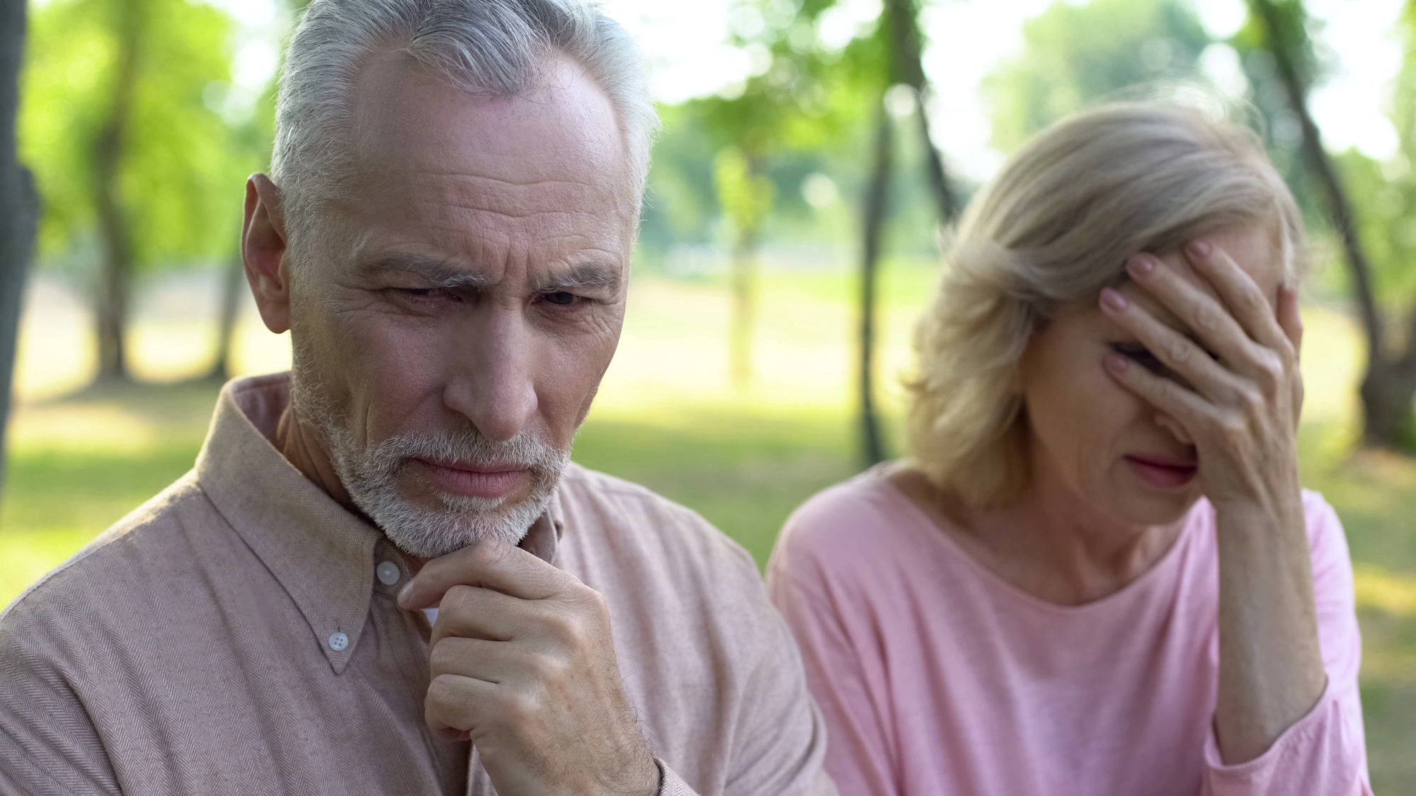 An elderly man and woman are outdoors. The man looks thoughtful, resting his chin on his hand, while the woman covers her face with her hand, appearing distressed. They are wearing casual clothing, and trees are visible in the background.