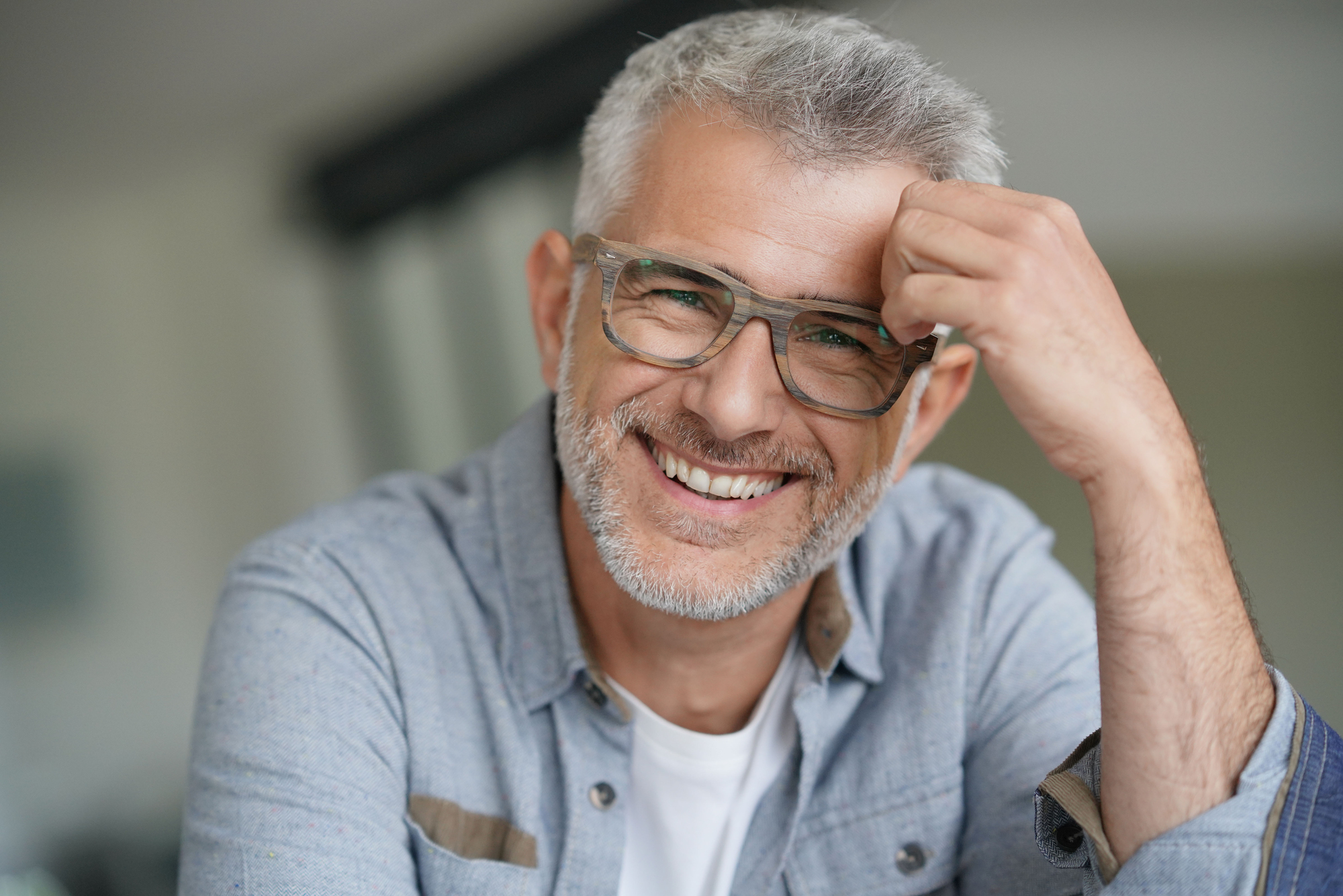 A smiling man with gray hair and glasses wearing a blue shirt sits indoors, resting his head on his hand.