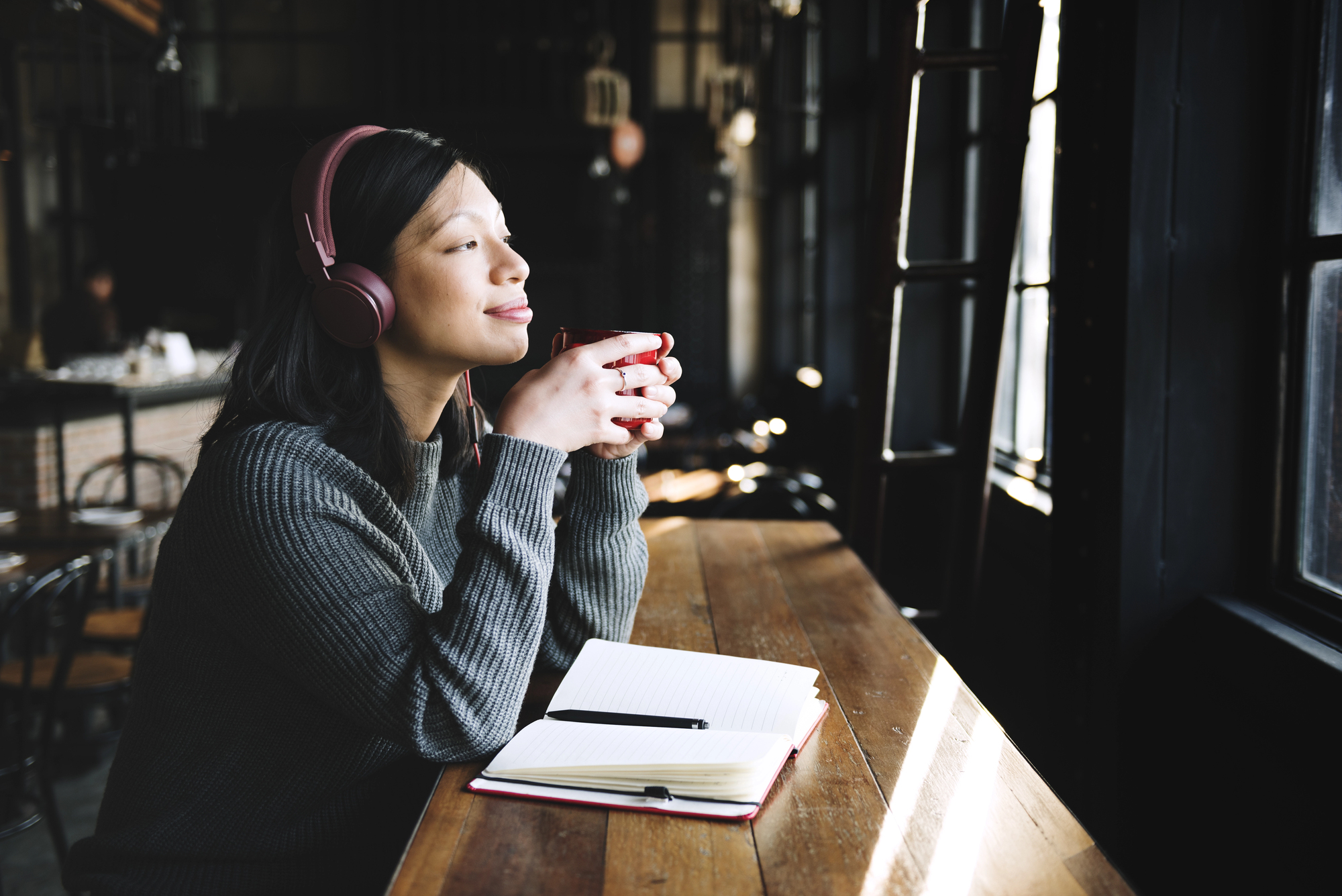 A person sits at a wooden table by a window, wearing headphones and holding a coffee cup, looking relaxed and content. An open notebook with a pen lies in front of them, and the background shows a cozy, dimly lit cafe.