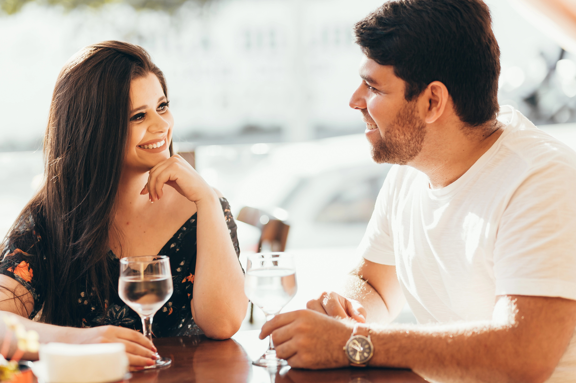 A woman and a man sit at a table outdoors, smiling at each other. They each have a glass of water in front of them. The sun is shining brightly, creating a warm and cheerful atmosphere.