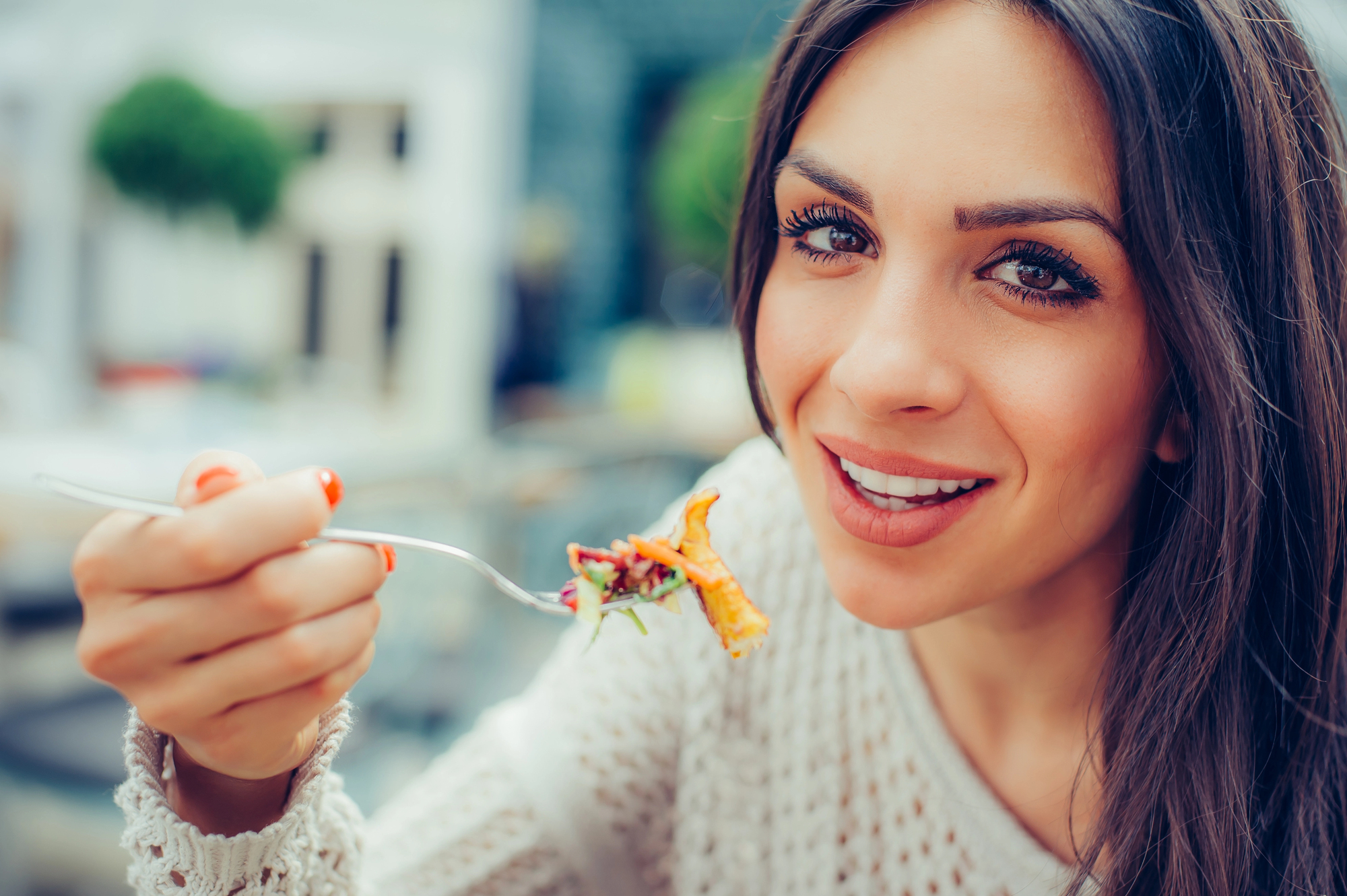 A smiling person with long hair holds a fork with food close to their mouth, sitting at an outdoor cafe. They're wearing a knitted sweater, and the background is blurred, featuring greenery and soft natural lighting.