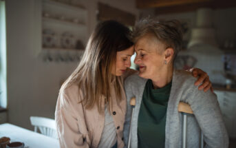 A younger woman and an older woman are embracing warmly in a cozy kitchen. The older woman, who uses crutches, has short gray hair and smiles gently. The younger woman, with long brown hair, leans her forehead against the older woman's, showing care and affection.