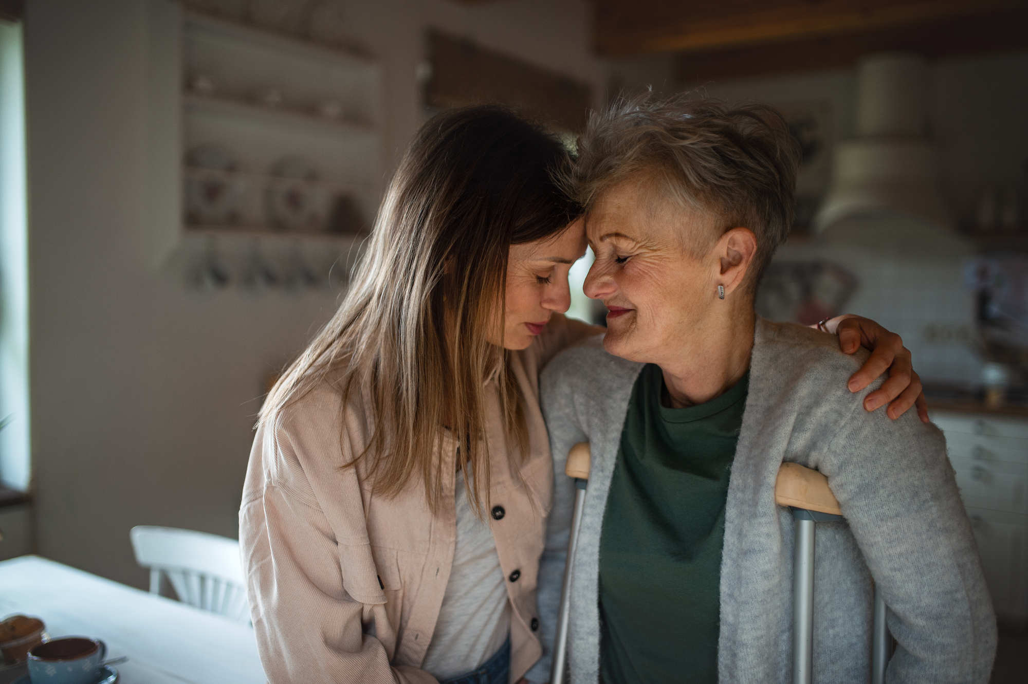 A younger woman and an older woman are embracing warmly in a cozy kitchen. The older woman, who uses crutches, has short gray hair and smiles gently. The younger woman, with long brown hair, leans her forehead against the older woman's, showing care and affection.