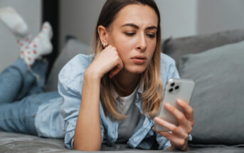 A woman with long hair and a casual outfit lies on a couch, looking intently at a smartphone in her hand. She rests her chin on her other hand, appearing focused and thoughtful.