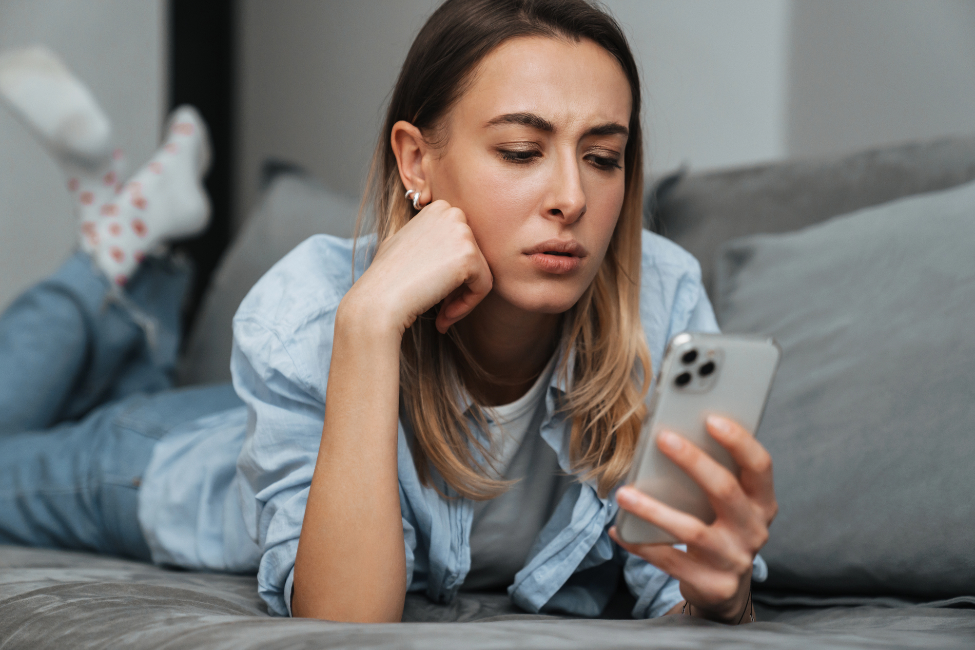 A woman with long hair and a casual outfit lies on a couch, looking intently at a smartphone in her hand. She rests her chin on her other hand, appearing focused and thoughtful.