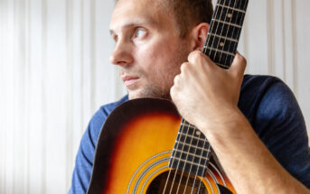 A man in a blue shirt holding an acoustic guitar close to his face, gazing pensively to the side. The guitar has a sunburst finish, and the background is a light-colored wall with vertical paneling.