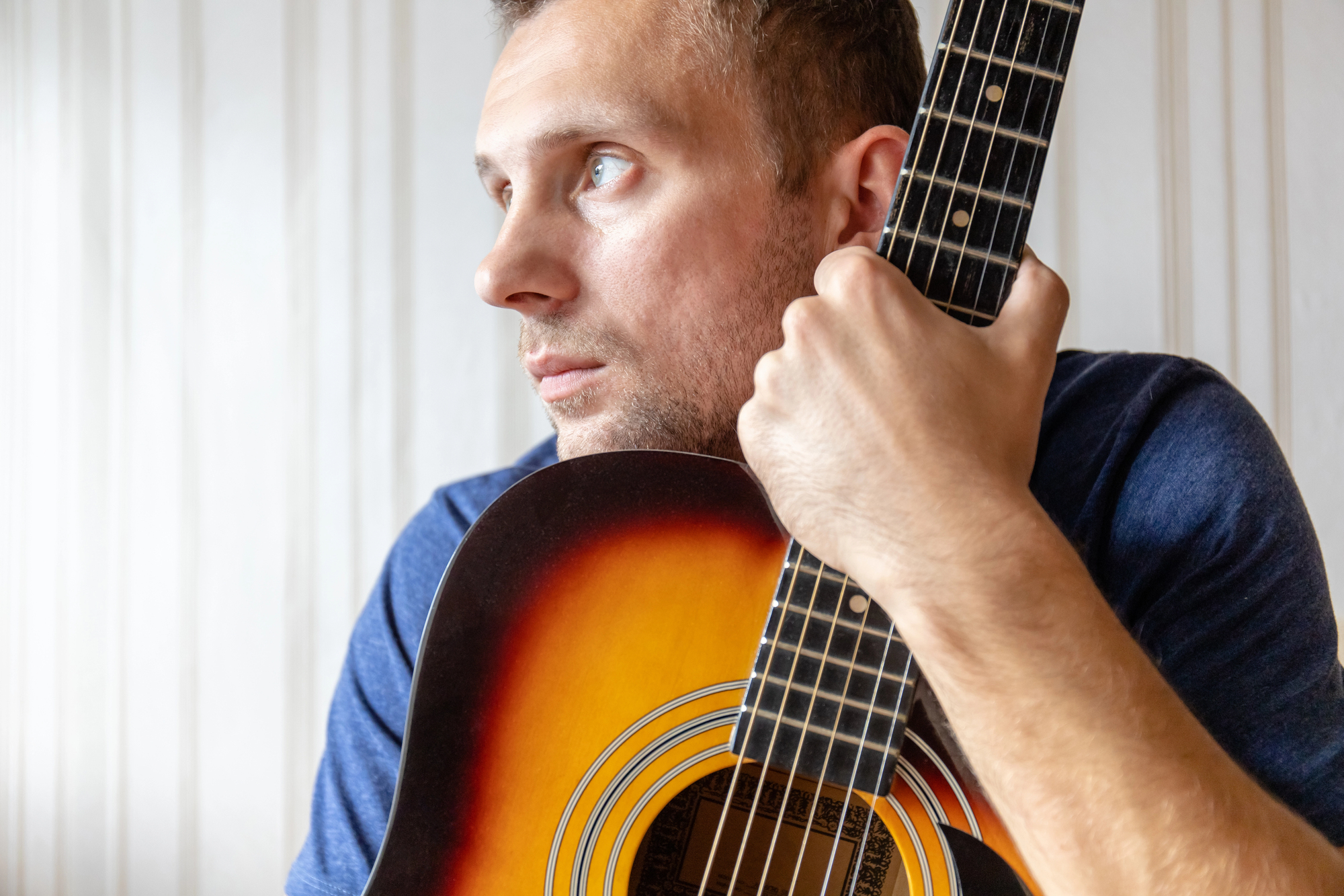 A man in a blue shirt holding an acoustic guitar close to his face, gazing pensively to the side. The guitar has a sunburst finish, and the background is a light-colored wall with vertical paneling.