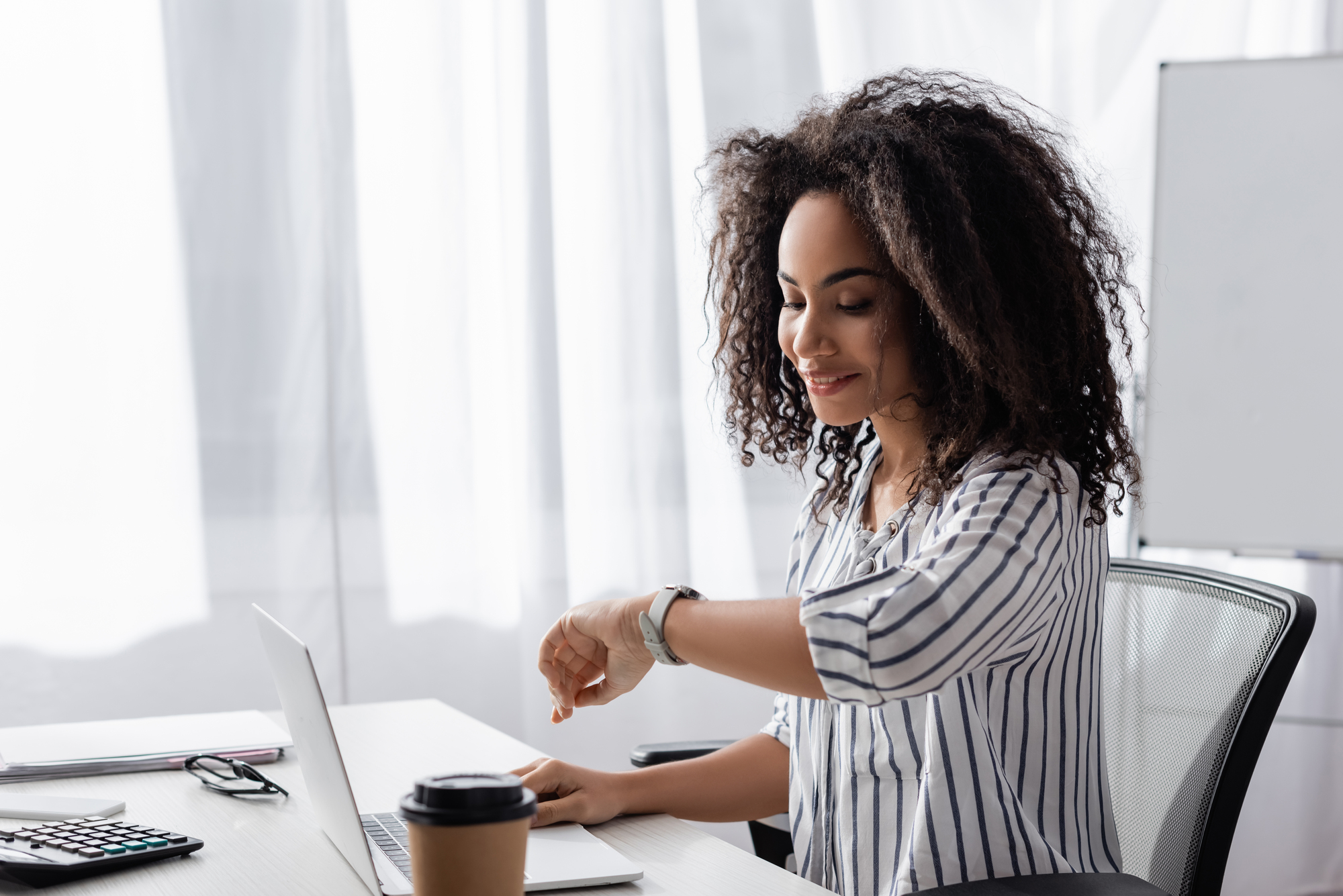 A woman with curly hair sits at a desk, looking at her smartwatch. She is in a striped shirt and smiles slightly, working on a laptop. A coffee cup is on the desk, and a bright window is in the background.