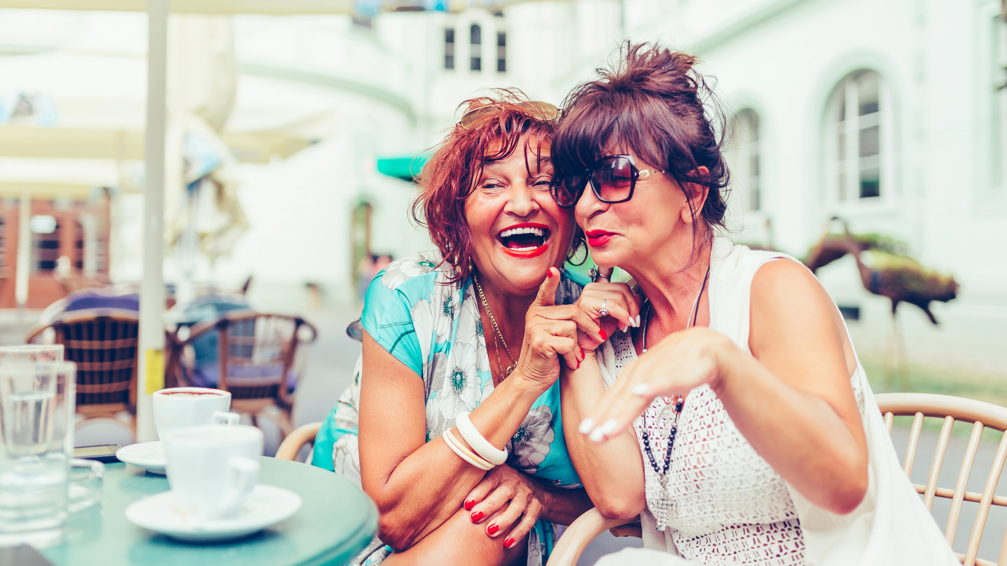 Two women sitting at an outdoor café, smiling and appearing to share a joke. One woman is wearing sunglasses, and the other is laughing joyfully. Coffee cups are on the table in front of them. The background shows a building and patio furniture.