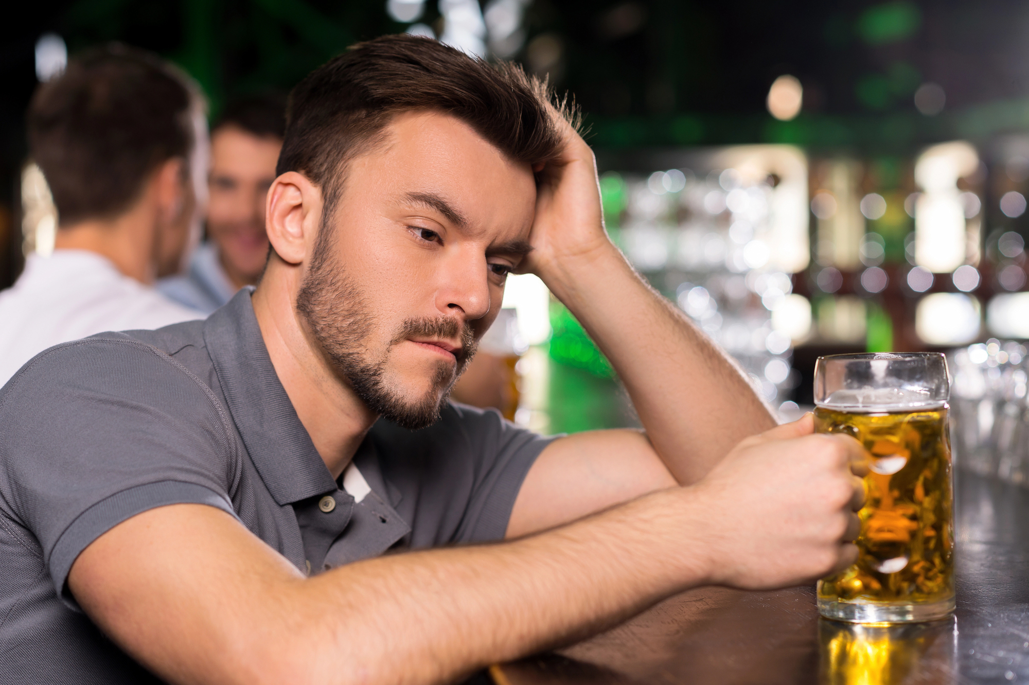 A man sitting at a bar, looking contemplative while holding a mug of beer. Other people are blurred in the background.