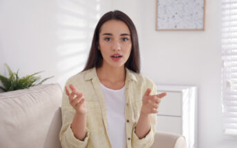 A woman with long dark hair is sitting on a beige sofa in a bright room, wearing a yellow striped shirt and white top. She appears to be speaking, with her hands slightly raised. Sunlight filters through a window with blinds, casting shadows.