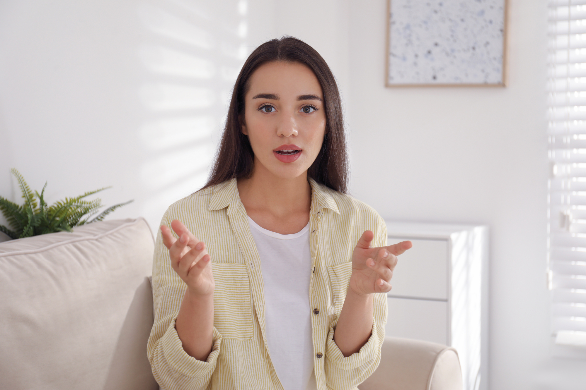 A woman with long dark hair is sitting on a beige sofa in a bright room, wearing a yellow striped shirt and white top. She appears to be speaking, with her hands slightly raised. Sunlight filters through a window with blinds, casting shadows.