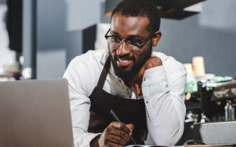 A man wearing glasses and an apron is smiling while looking at a laptop. He is holding a pen and writing in a notebook in front of him. There is a coffee machine in the background.